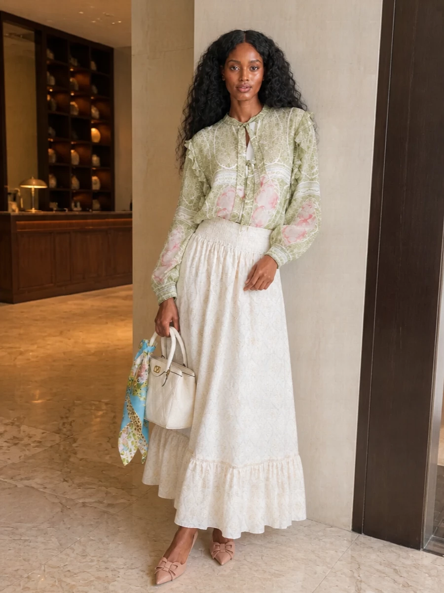 Sage florals and whisper-light ivory create a romantic, softly bohemian ensemble that feels poised yet relaxed. The fluid maxi skirt echoes the blouse’s movement, while nude-blush heels and an ivory top-handle keep the palette light and refined. A silk carré adds a subtle wash of colour and texture, tying the whole composition together with quiet charm.