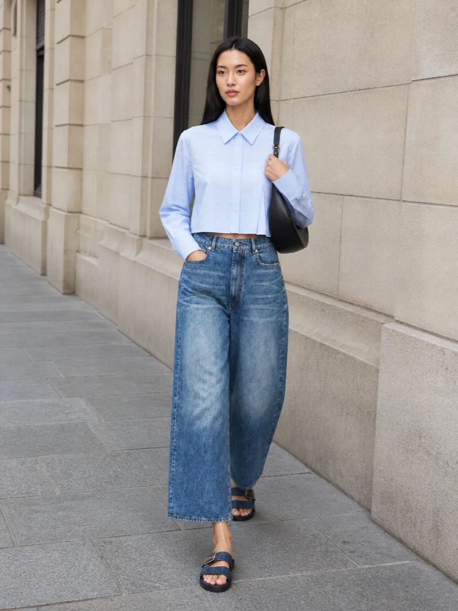 Cropped architectural shirting in pale blue sits precisely on the high-rise, culotte-like jeans, keeping the volume focused in the leg while the torso stays clean and defined. Denim slides underline the relaxed, SS ease, and a sculpted black crescent shoulder bag adds a quiet, graphic finish that keeps the whole look minimal, directional and city-ready.