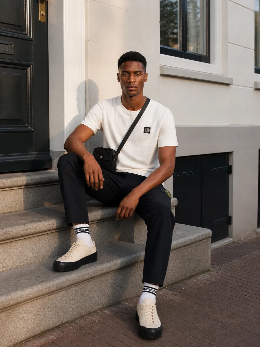Off-white Stone Island jersey on top of deep navy straight-leg chinos sets a clean, low-contrast base that still feels graphic. Cream and sand runners keep everything light at ground level, while the compact black crossbody adds a technical note without interrupting the lines. Collegiate striped socks are the only hint of color, connecting the neutrals and keeping the whole outfit quietly sporty and considered for warm days in the city.
