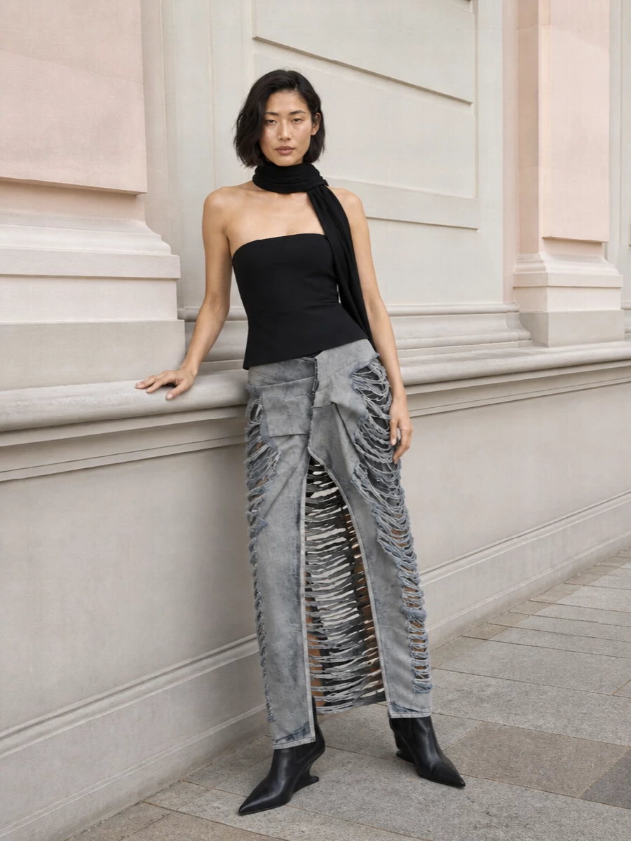 A high‑waisted, laddered grey denim column meets a stark black strapless top for a severe, night‑driven silhouette that feels almost carved out of shadow. Sculptural black ankle boots and a compact crescent bag keep the lines clean and vertical, letting the negative space in the skirt do the talking. The effect is pure Rick energy: brutal, elongated and unapologetically after‑dark.