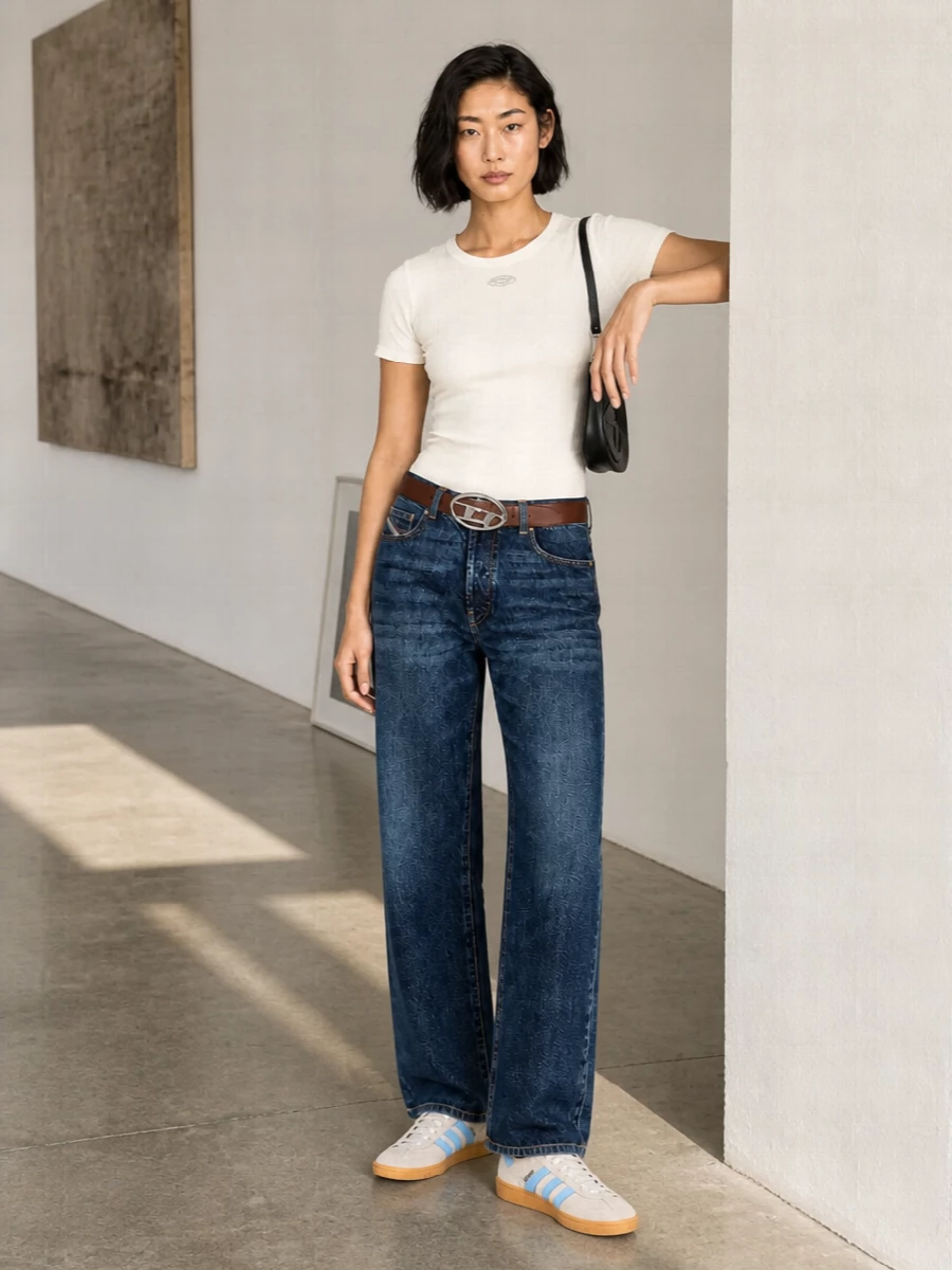 Dark straight-leg Diesel denim meets a shrunken ivory tee for a clean, low-key streetwear take that still feels intentional. Retro beige Adidas sneakers and a black Diesel shoulder bag keep it grounded, with a tan sculptural belt tying together the warm stitching and gum tones. The result is relaxed, graphic and easy to wear from city errands to late casual dinners.