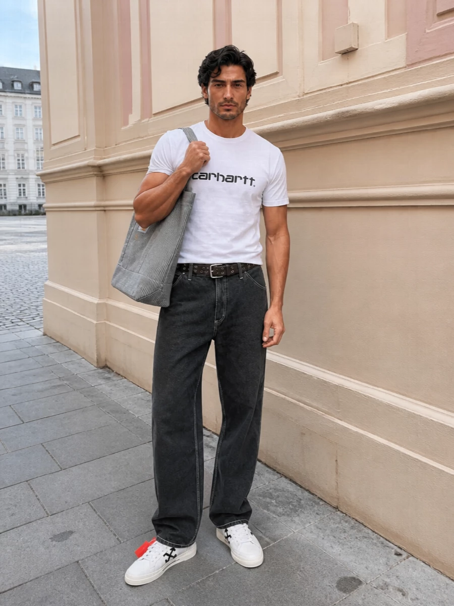 A white Carhartt logo tee sits against washed charcoal workwear trousers for a sharp, monochrome take on skate-inspired streetwear. Off-White court sneakers and a striped Carhartt tote keep the mood clean but graphic, while the grommeted belt adds just enough industrial detail at the waist. It is an easy city uniform that feels considered without trying too hard.