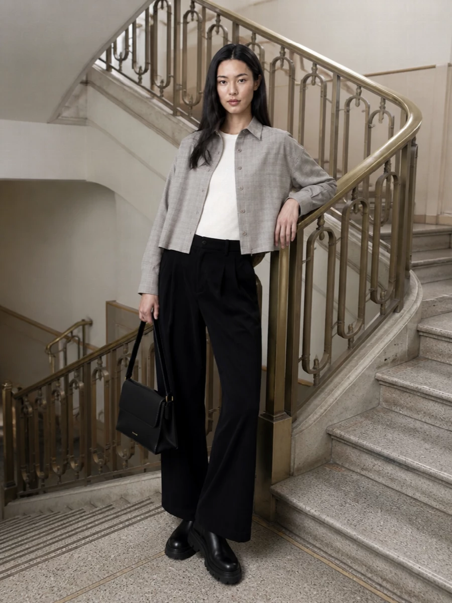 I’m styling the cropped checked shacket with a simple tee and tailored wide-leg trousers rather than a dress to preserve its easy, borrowed-from-the-boys energy. The long, fluid black cord anchors the boxy, cropped outerwear, while black leather boots and a structured shoulder bag keep everything sharp but relaxed enough for all-day city wear. Soft taupe and grey checks sit quietly against the monochrome base, so the whole outfit reads modern and intentional without trying too hard.