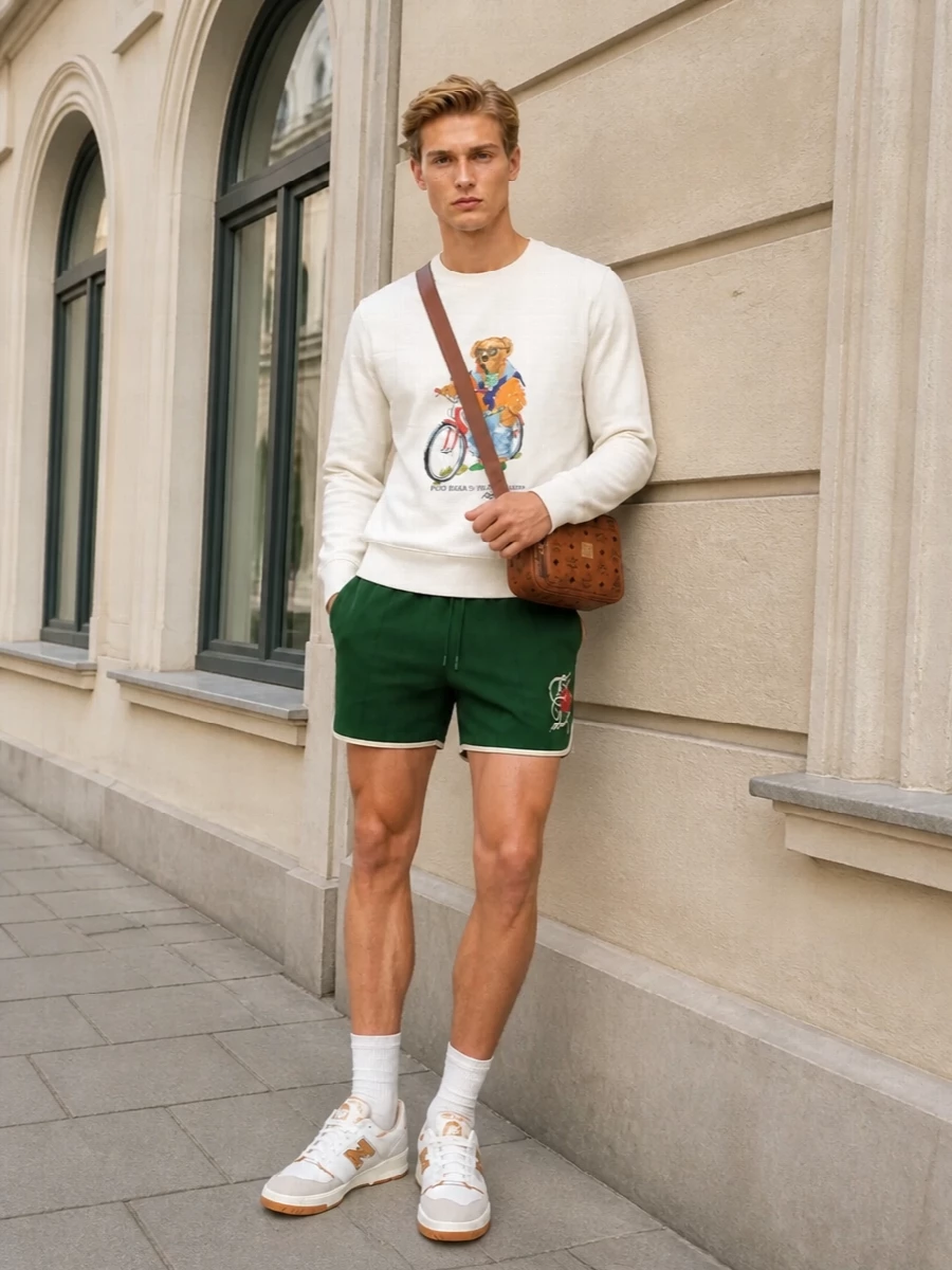 A cream Polo Bear sweatshirt meets rich forest-green sport shorts for a nostalgic campus-sport mood that still feels fresh for warm days. Retro-influenced sneakers pick up the orange from the graphic, while the cognac crossbody leans into classic, slightly polished preppy energy without losing the relaxed vibe.