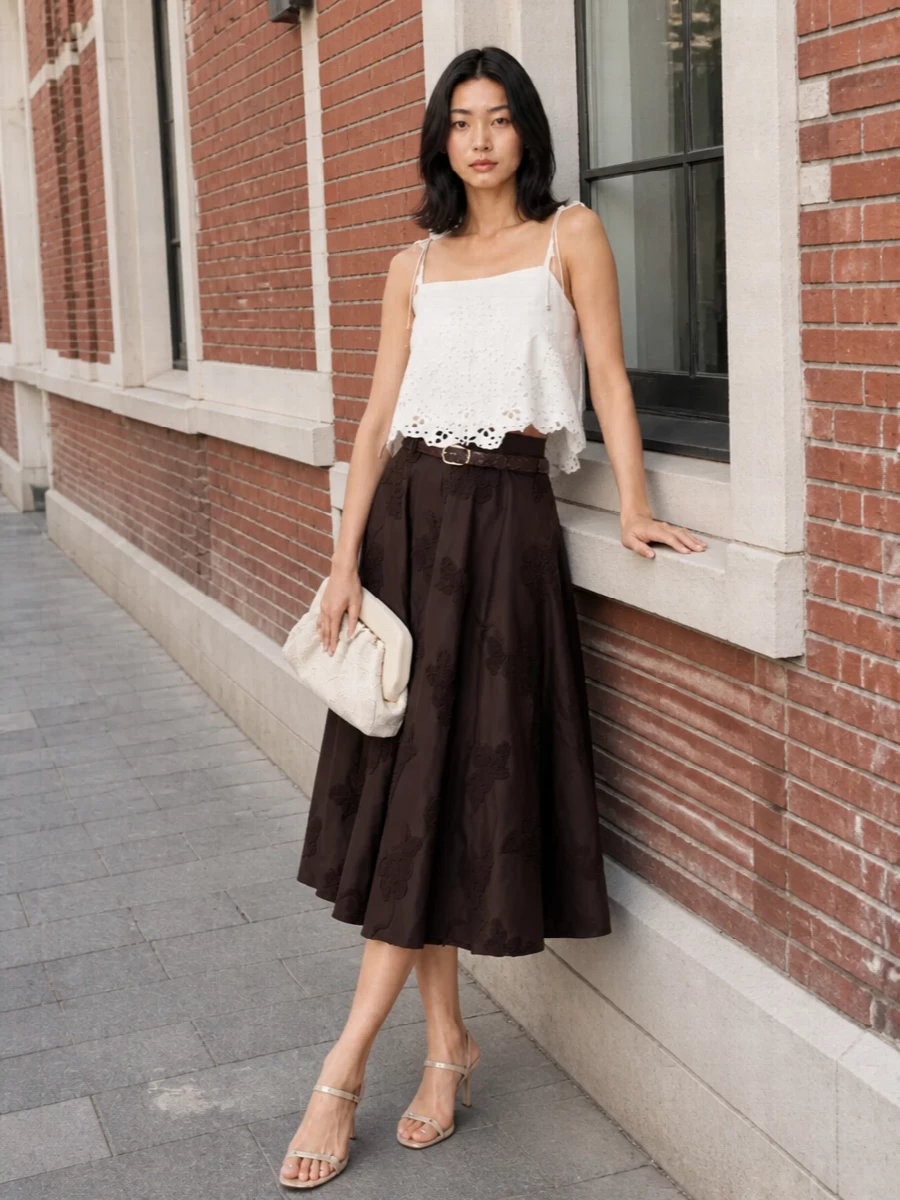 A deep chocolate embroidered midi skirt is softened with an ivory broderie camisole for a romantic, quietly polished dinner ensemble. Champagne ankle‑strap sandals and an ivory framed clutch keep the palette luminous, while a slim chocolate braided belt subtly defines the waist and echoes the skirt’s texture. The result feels feminine and composed, ideal for warm‑weather cocktails or intimate restaurant evenings.