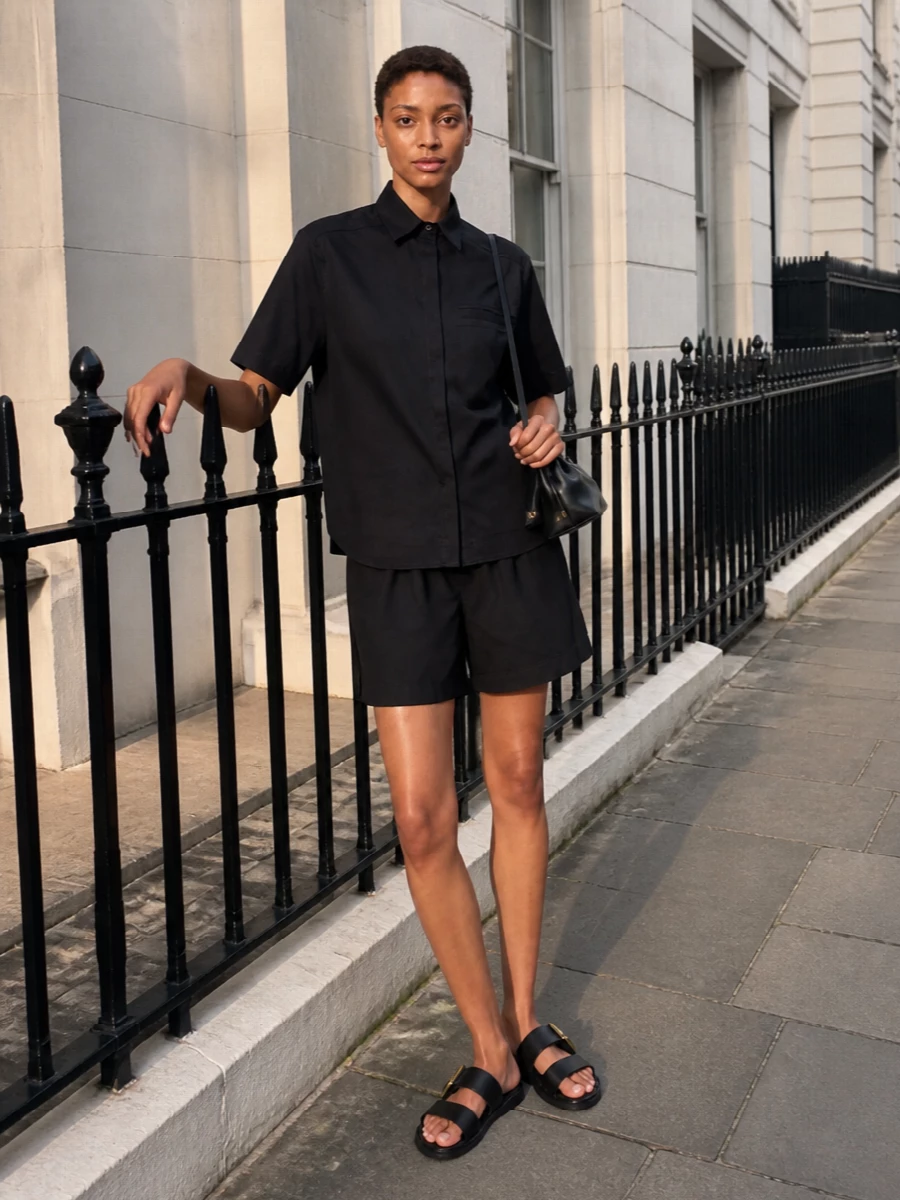 A black-on-black pairing keeps the Lemaire boxer short feeling intentional, not casual, with the crisp short-sleeve shirt giving it a quietly tailored frame. Flat black slides and a compact black pouch keep the silhouette low and unfussy, ideal for warm days in the city when you still want the outfit to read considered. The effect is relaxed, minimal and quietly sensual, all in a tight, understated palette.