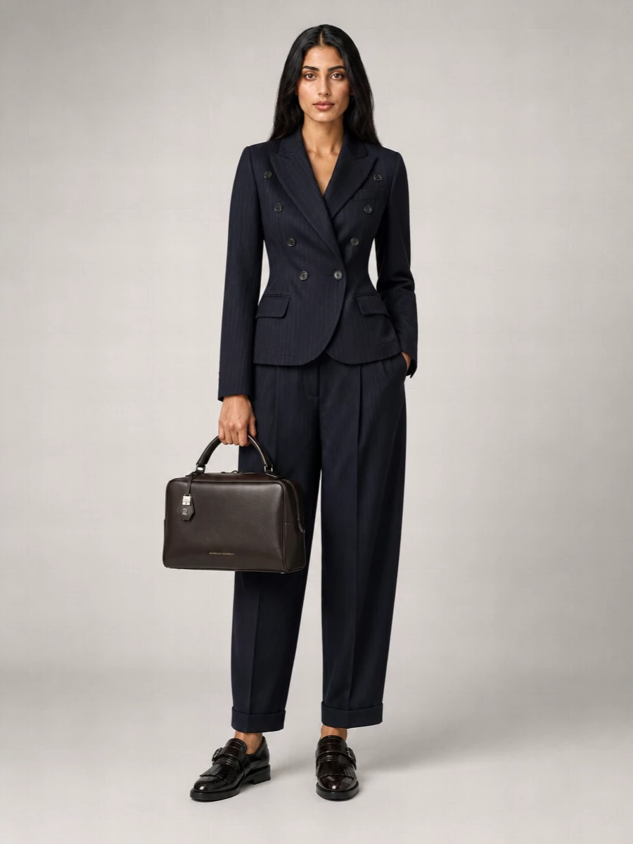 Navy-on-navy tailoring keeps the Vivienne Westwood blazer and pleated trouser line sharp, with the high waist and subtle cuff adding that quietly preppy twist. Grounded by a heritage monk shoe and a structured espresso top-handle bag, this is precise desk-to-dinner suiting that feels composed without being severe.