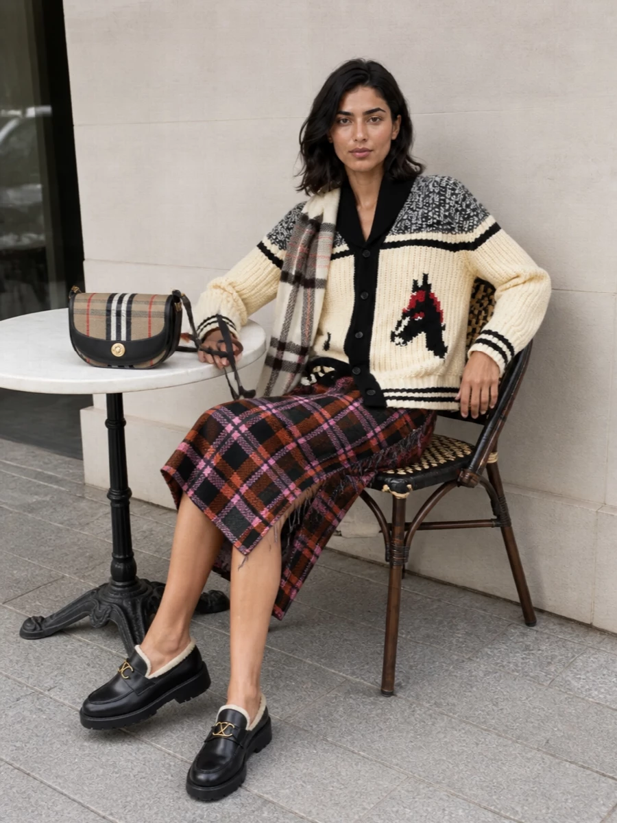 A chunky varsity cardigan tops a graphic plaid knit skirt for a collegiate mix of texture and pattern that still reads intentional. Solid black lug-sole loafers and a structured heritage-check shoulder bag ground the volume and color, while a soft cream check scarf adds winter warmth and a quiet nod to the prep references.