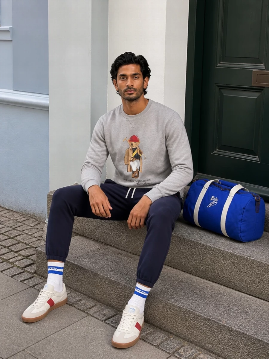 A heather-grey Polo Bear sweatshirt pairs with clean navy tapered joggers for a classic campus-inspired sport mood. Off-white Adidas sneakers with a red stripe echo the graphic, while the royal-blue Polo duffle leans into that collegiate energy for gym runs and weekend travel. Soft striped socks finish things off with a small, considered nod to preppy detail when the jogger cuffs sit just at the ankle.