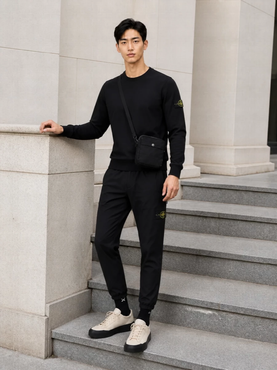 Black Stone Island track pants with the matching black logo sweatshirt create a tight, technical base where the neon patches do the talking. Cream and sand sneakers lift the darkness just enough, and a compact black crossbody plus crisp black socks keep everything streamlined for city movement. It is a clean, athletic streetwear ensemble built for travel, errands and low-key weekends on repeat.