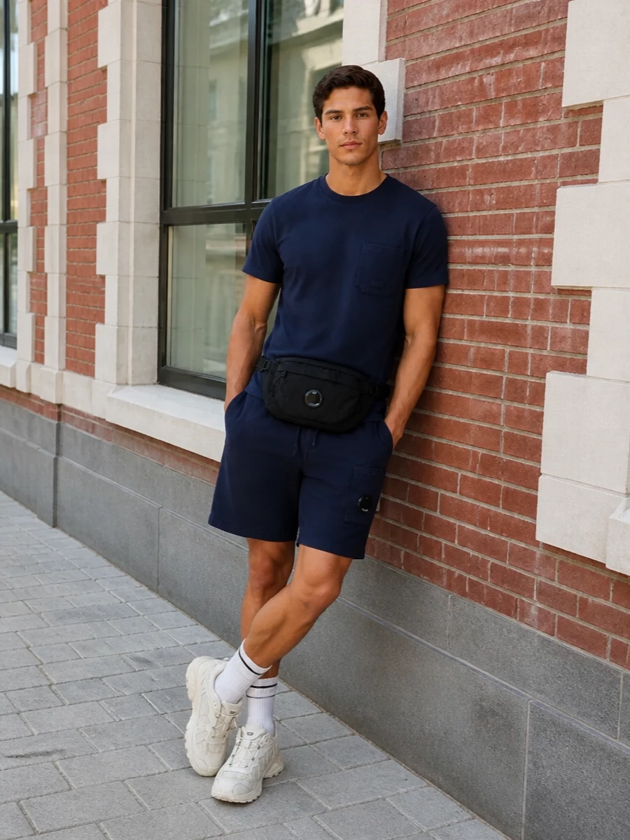 Navy-on-navy C.P. Company anchors this with a clean tee and technical shorts, keeping the silhouette sharp but easy for travel or city days. Off-white trail runners and matching sock accents break up the darkness just enough, adding a fresh, performance edge that still feels quietly refined. The compact belt bag finishes it with hands-free utility and a repeat of that signature lens detail.