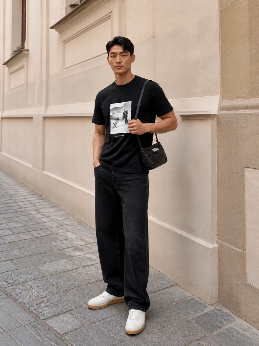 A black cinematic tee sits against relaxed washed-black denim for a monochrome street uniform that feels precise rather than stark. Off-white gum-sole sneakers break the darkness just enough, and a padded Margiela mini bag plus graphic socks keep the branding tight and considered. Wear the denim long over the sneakers so the line stays easy and unfussy.