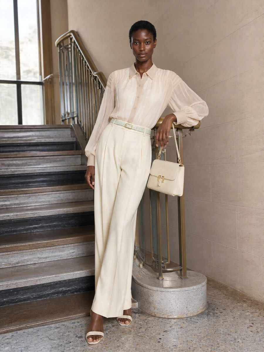 Buttercream tailoring stays softly luminous with Alberta Ferretti’s sheer ivory blouse tucked into the high-rise wide-leg trousers, creating a statuesque yet weightless column. Cream ankle-strap sandals and a structured cream top-handle bag keep the line clean and elongated, while the slim scalloped belt adds a discreet touch of definition and color without disturbing the quiet luxury mood.