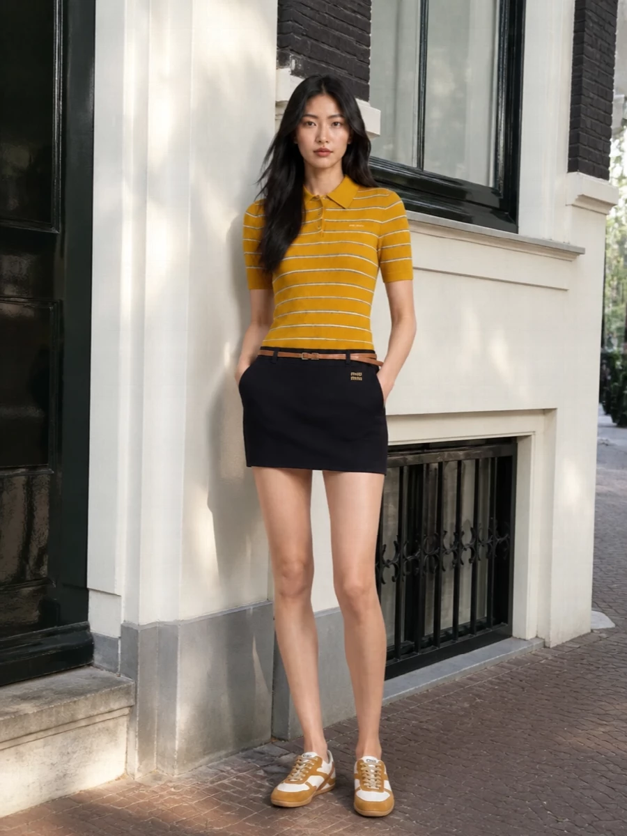Sunlit mustard and deep navy play up the Miu Miu polo’s collegiate attitude, grounded by a clean, uniform-feel mini skirt. Retro trainers keep it relaxed and agile, while the tonal suede top-handle and slim chain-detailed belt make the whole outfit feel considered rather than costume. It is polished campus energy for city days, with just enough Y2K to stay fun.