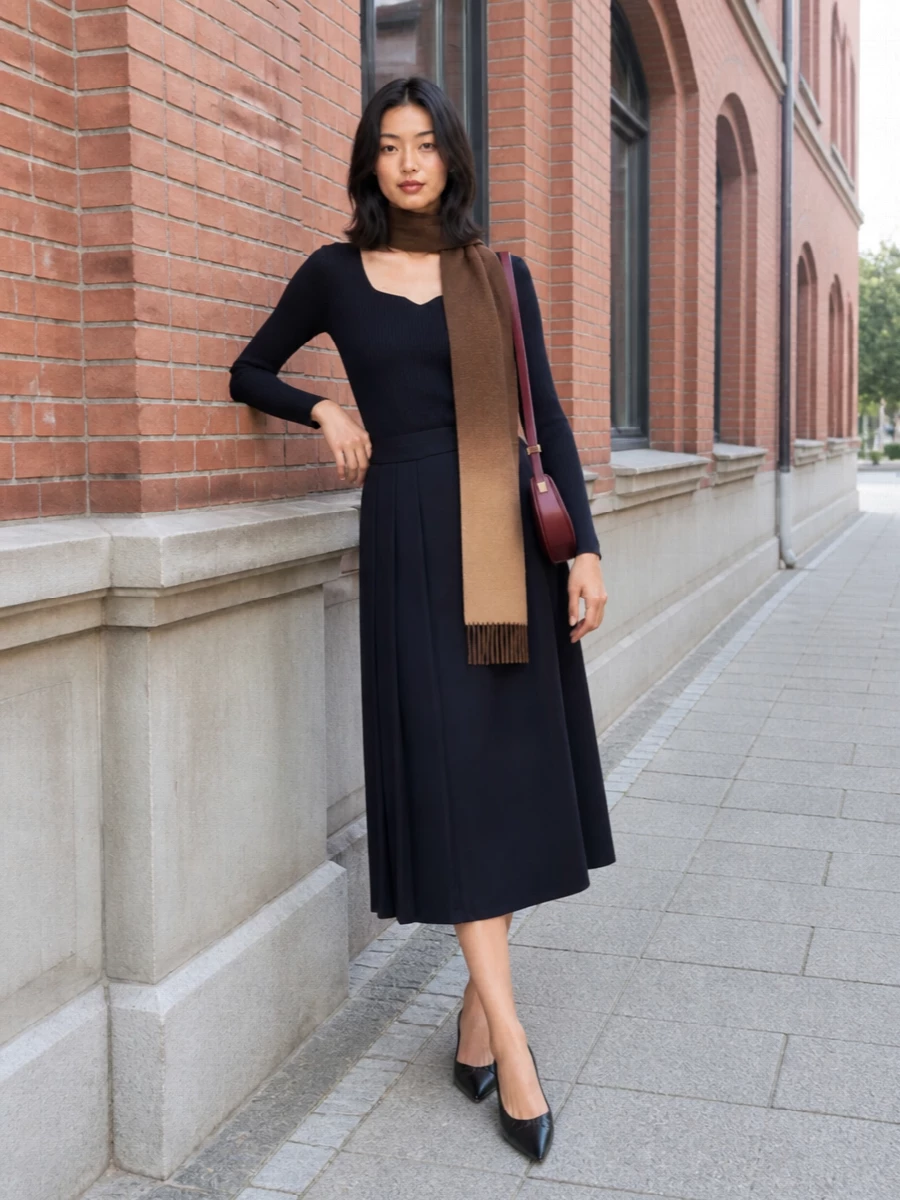 Deep navy rib and pleats create a long, clean column, lightly broken by a pointed black kitten heel that keeps everything sharp but gentle. A small burgundy shoulder bag and warm ombré scarf introduce soft color without disturbing the minimalist palette, so the outfit still feels quietly romantic and desk-to-dinner ready. The result is a slim, fluid silhouette that reads considered rather than severe.