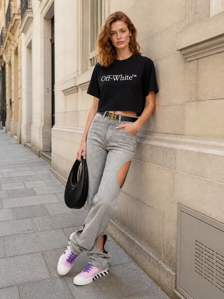 This look leans fully into Off-White’s graphic streetwear codes: the cropped logo tee sits over slouchy grey cut-out denim for a bold, leggy silhouette with attitude. Pastel-pink low-top sneakers keep it casual and wearable, while the sculptural black Coperni bag and a clean logo belt add just enough polish to take the outfit from daytime city roaming to after-dark plans without losing its edge.