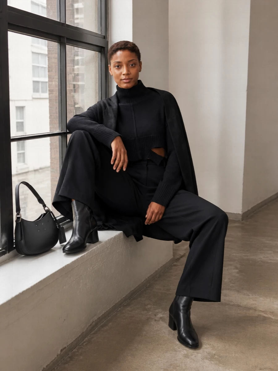 A cropped, deconstructed knit sits against long, high-waisted wide-leg trousers for a sharp Margiela column in pure black. Sculptural ankle boots and a curved shoulder bag keep the mood minimal yet precise, with an oversized tonal wrap adding depth rather than distraction. It is a quietly directional streetwear-meets-tailoring uniform for cold city days and late gallery nights.