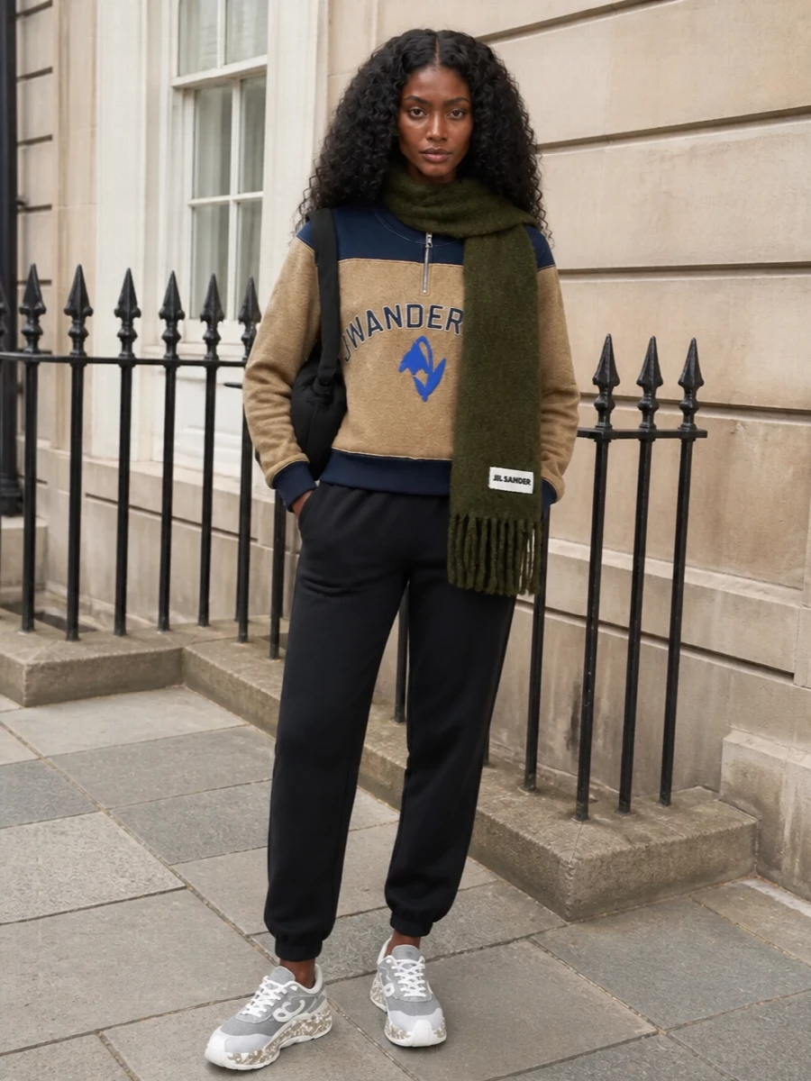 A camel and navy JW Anderson sweatshirt meets precision-cut black Jil Sander joggers for a sport-inflected city uniform that feels relaxed but highly considered. Chunky grey Loewe sneakers keep the energy grounded and modern, while a knotted black shoulder bag and an olive scarf add quiet texture and depth without stealing focus from the graphic top.