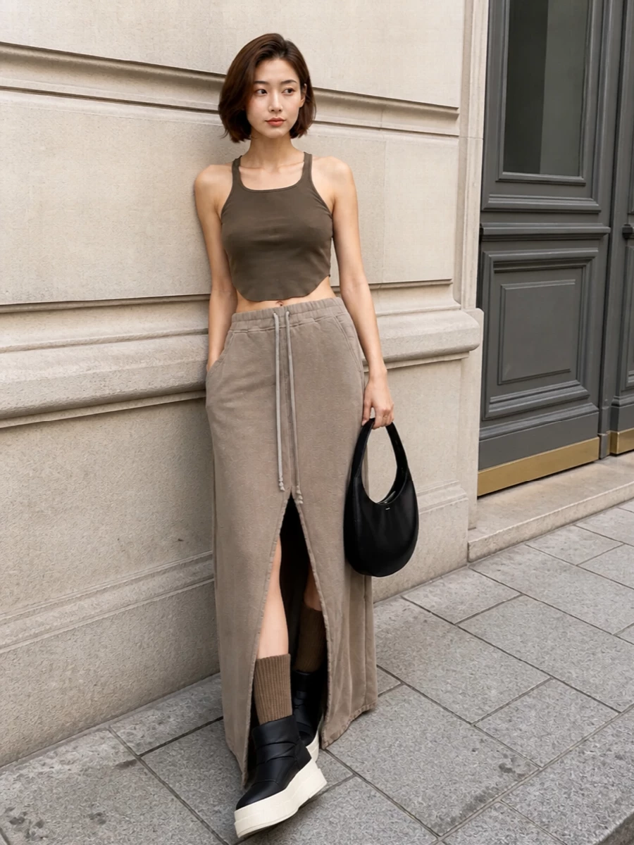 Mud-brown DRKSHDW tank tucks into the taupe drawstring maxi, creating a long, tonal column that feels both relaxed and subversively athletic. Chunky padded platform boots ground the softness with graphic black, while a sculpted black Coperni hobo bag introduces a clean, futuristic edge that keeps the look club-ready yet understated. Taupe knee-high socks disappear into the palette, sharpening the elongated, underworld-sport silhouette without distracting from the minimal lines.