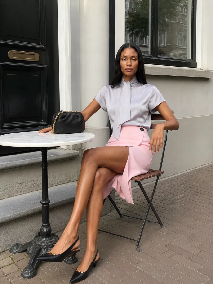 Soft blush and dove grey create a calm, polished canvas, with the satin tie-neck adding just enough movement against the skirt’s asymmetric wrap. Black accents in the shoes and clutch echo the skirt’s button, grounding the palette and keeping the ensemble city-ready for cocktails or discreet evening events.