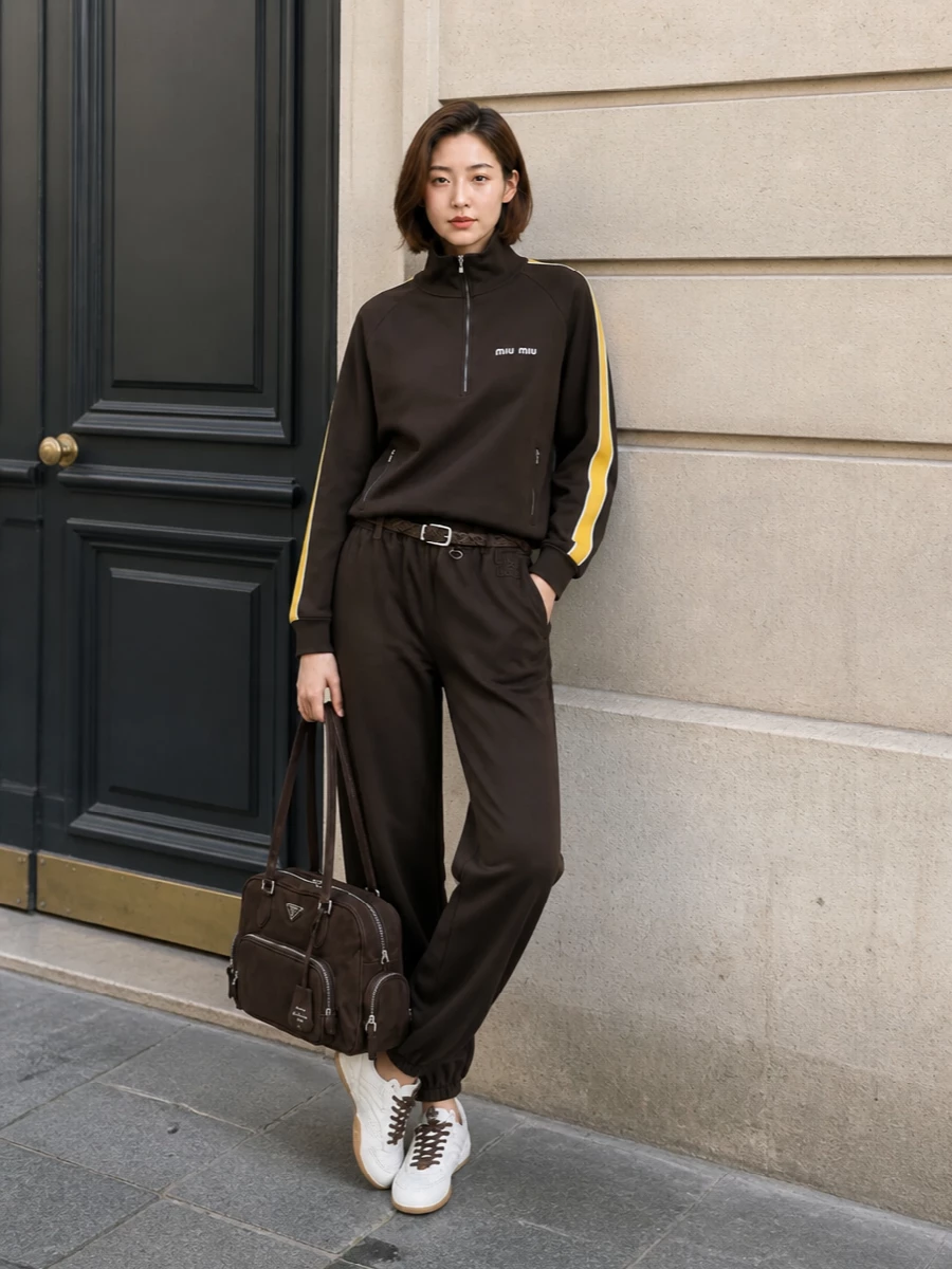 This look leans into a rich, chocolate monochrome, pairing Miu Miu’s elastic-cuff track pant with the matching-toned half‑zip for a streamlined, elevated athleisure set. Off‑white-and-brown low-top sneakers keep the proportions light and agile, while the structured chocolate Prada tote and braided belt introduce quiet-luxury structure and texture without disturbing the sport-driven ease. The result is a polished, tonal city uniform that still feels legitimately athletic and SS-ready.