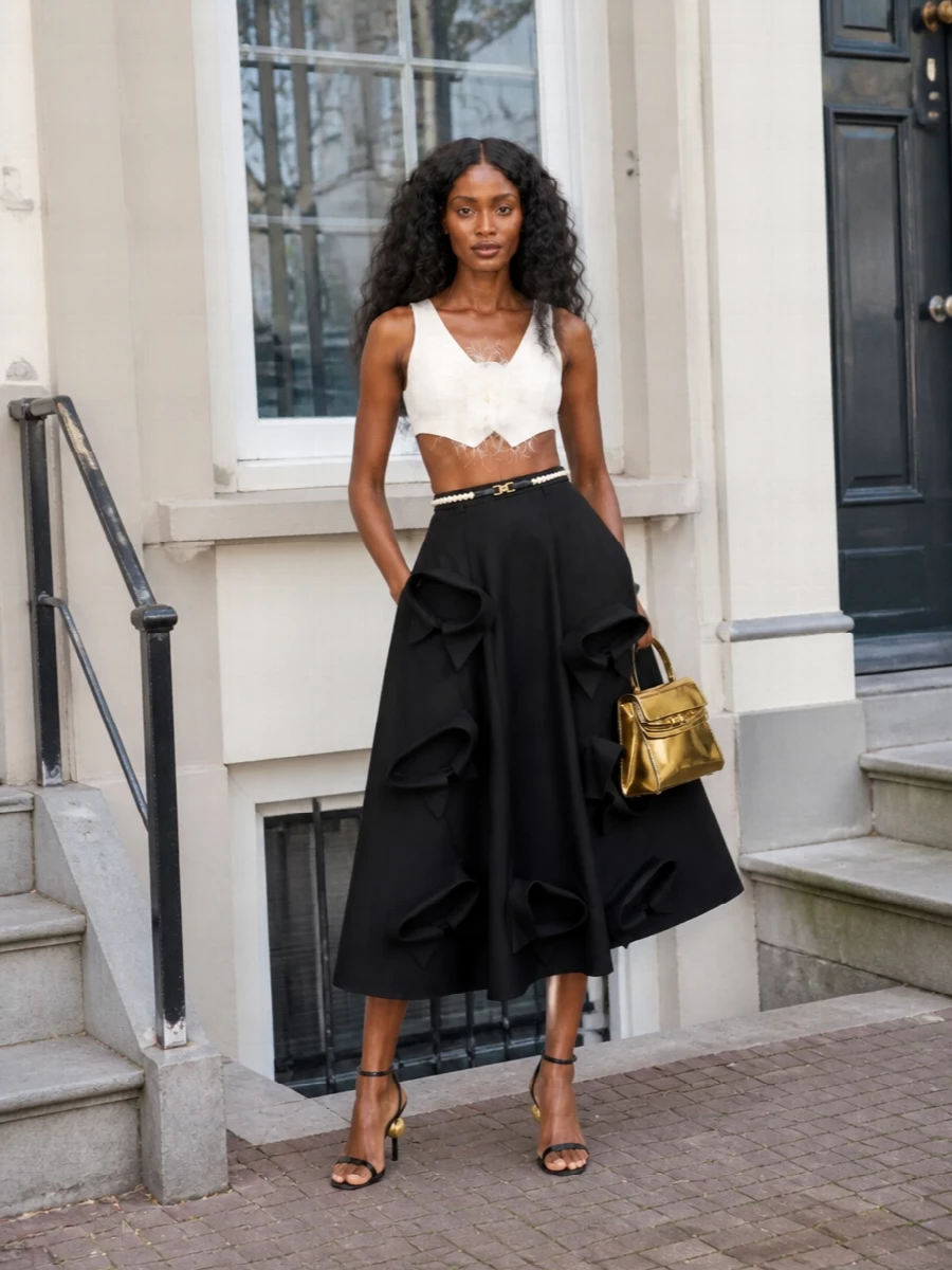 A feathered ivory bralette tops a sculptural black bow-appliqué midi, creating a romantic, Y2K-inflected silhouette that still reads sharp and polished. Sleek black and gold strappy sandals lengthen the line, while a pearl-detailed waist and gleaming gold top-handle bag pull the drama into a focused, evening-ready statement. The result is playful cocktail dressing with couture-level detail and a clean monochrome base.