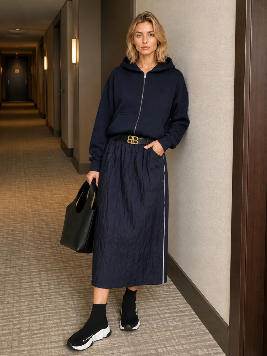 A navy technical skirt anchors a cocooning Balenciaga hoodie for a relaxed street uniform that still feels considered. Sock sneakers keep the lines fast and aerodynamic, while a sculptural black tote and slim logo belt bring just enough structure. The result is a monochrome, sport-inflected city outfit that reads directional without trying too hard.