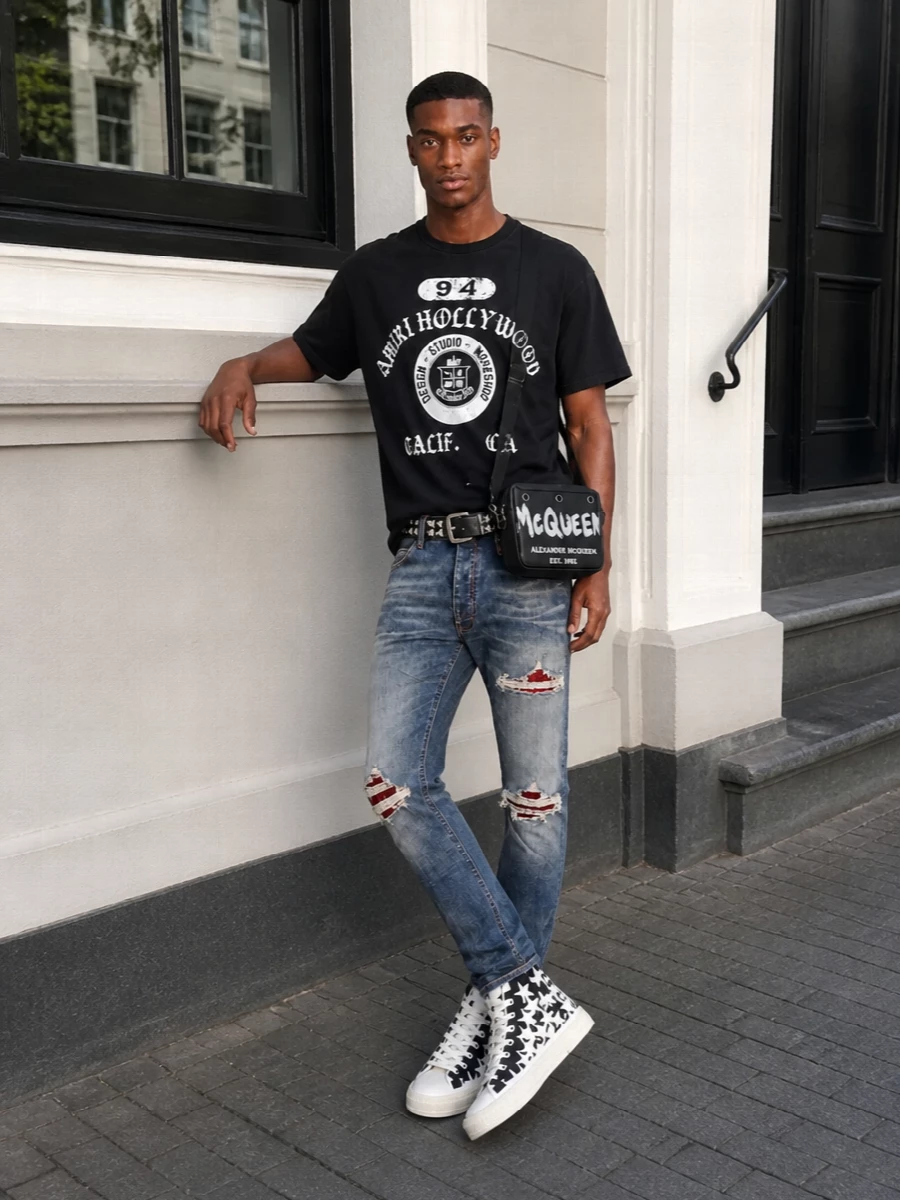 Amiri’s shredded striped denim sets a rock-stage foundation, met by a washed black Hollywood crest tee that keeps the energy graphic but grounded. Star-studded high-tops echo the jeans’ drama and add height, while the studded belt and bold McQueen crossbody pull the hardware story through the waist and shoulder. The result is a tightly edited, late-night LA street ensemble that feels intentional from hem to strap.