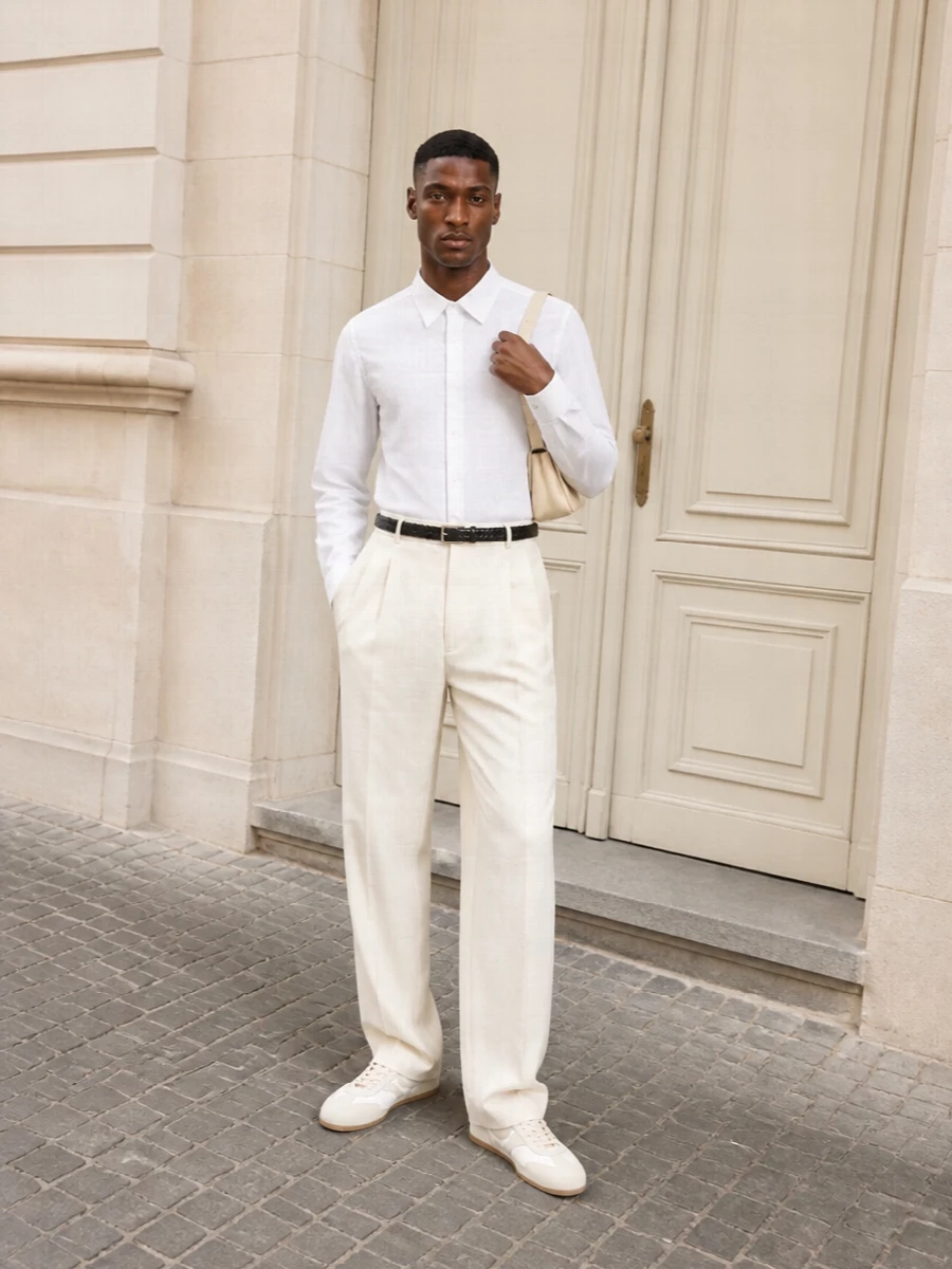 A crisp white shirt tucked into cream wide-leg pleats creates a clean, elongated line that feels calm yet precise. Soft off white runners and a buttercream shoulder bag keep everything in a tonal, modern neutral story. The slim braided belt subtly anchors the high rise so the volume reads intentional rather than oversized.