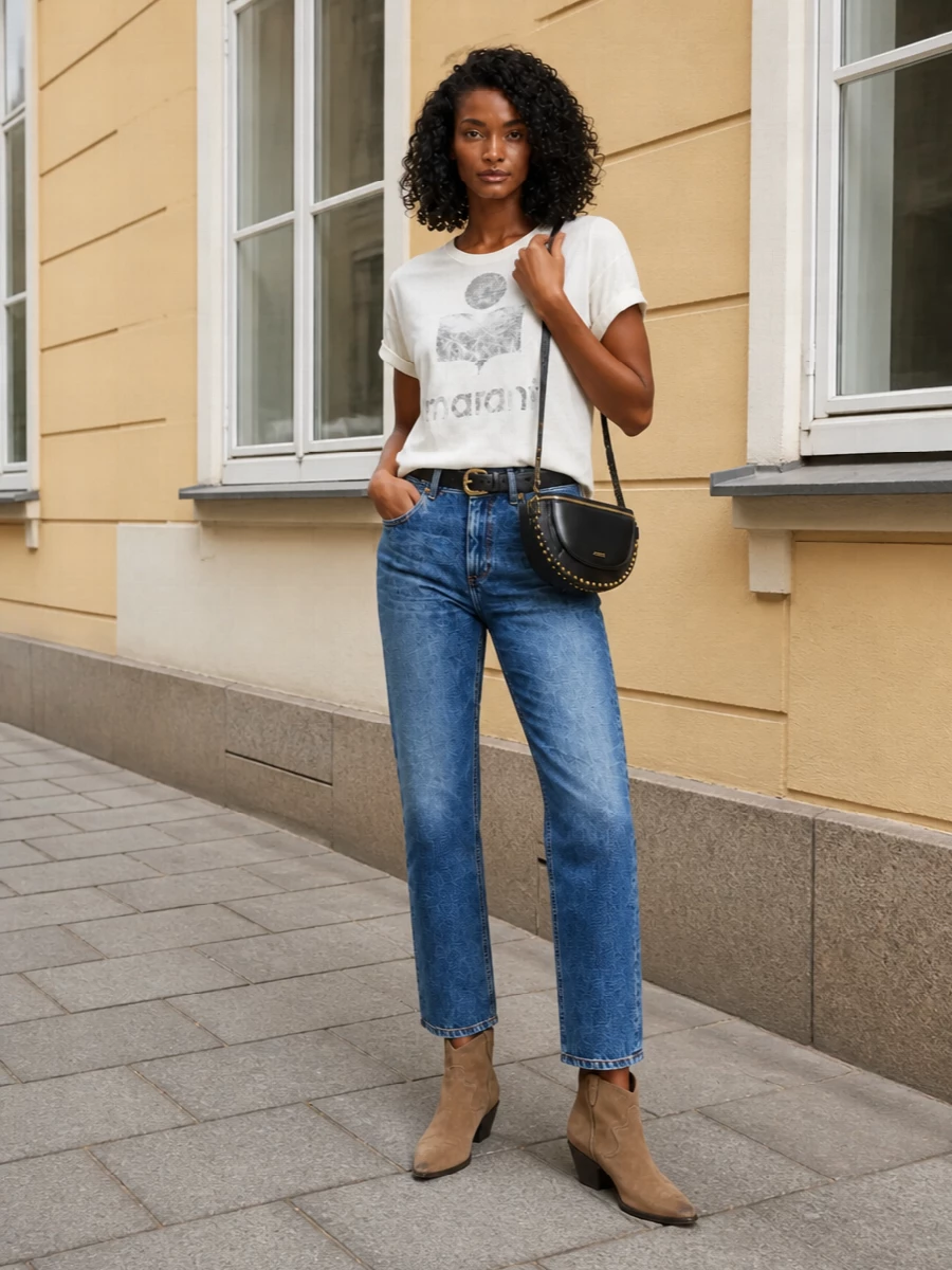 Soft white logo tee, gently faded straight-leg denim and taupe western boots create an easy, Paris-inflected streetwear uniform. A black studded saddle bag and slim belt add just enough polish while keeping the mood relaxed and low-key. The result is an all-season off-duty outfit that feels considered but never overworked.