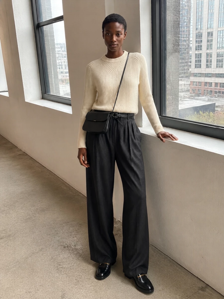 Charcoal wool trousers are paired with a softly structured buttercream ribbed knit, giving a calm, neutral contrast that still feels office ready. Black loafers and a streamlined black crossbody sharpen the finish, with a slim belt subtly defining the waist so the wide leg reads intentional rather than slouchy. The result is a quietly refined city uniform that moves easily from desk to low-key dinner.