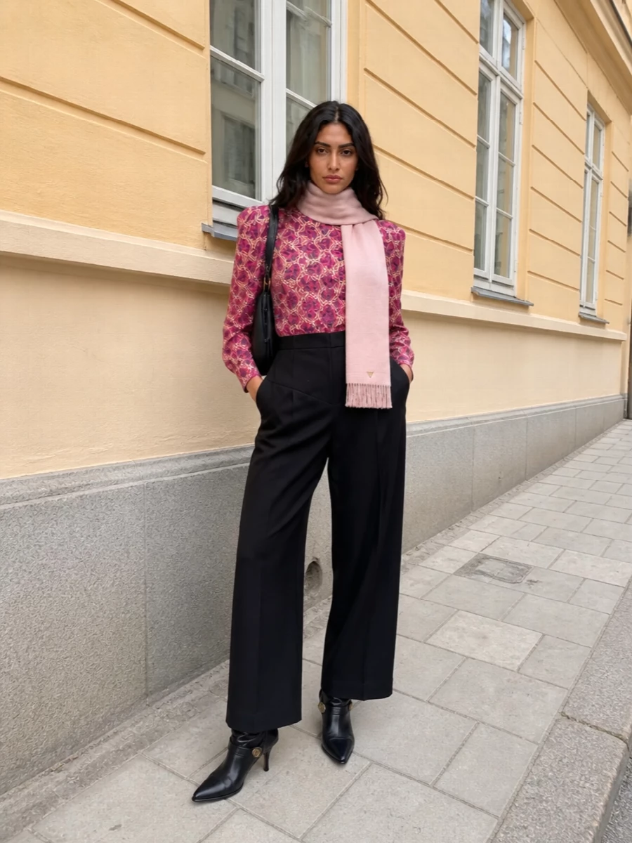 A vivid paisley blouse is sharpened by high-waisted black wide-leg trousers, creating a strong, sculpted line that moves easily from dinner to late drinks. Black stiletto boots and a structured leather shoulder bag keep the energy polished and modern, while a blush scarf adds a quiet, romantic softness against the saturated pink. The result is bohemian at heart but city-ready in its proportions and finish.
