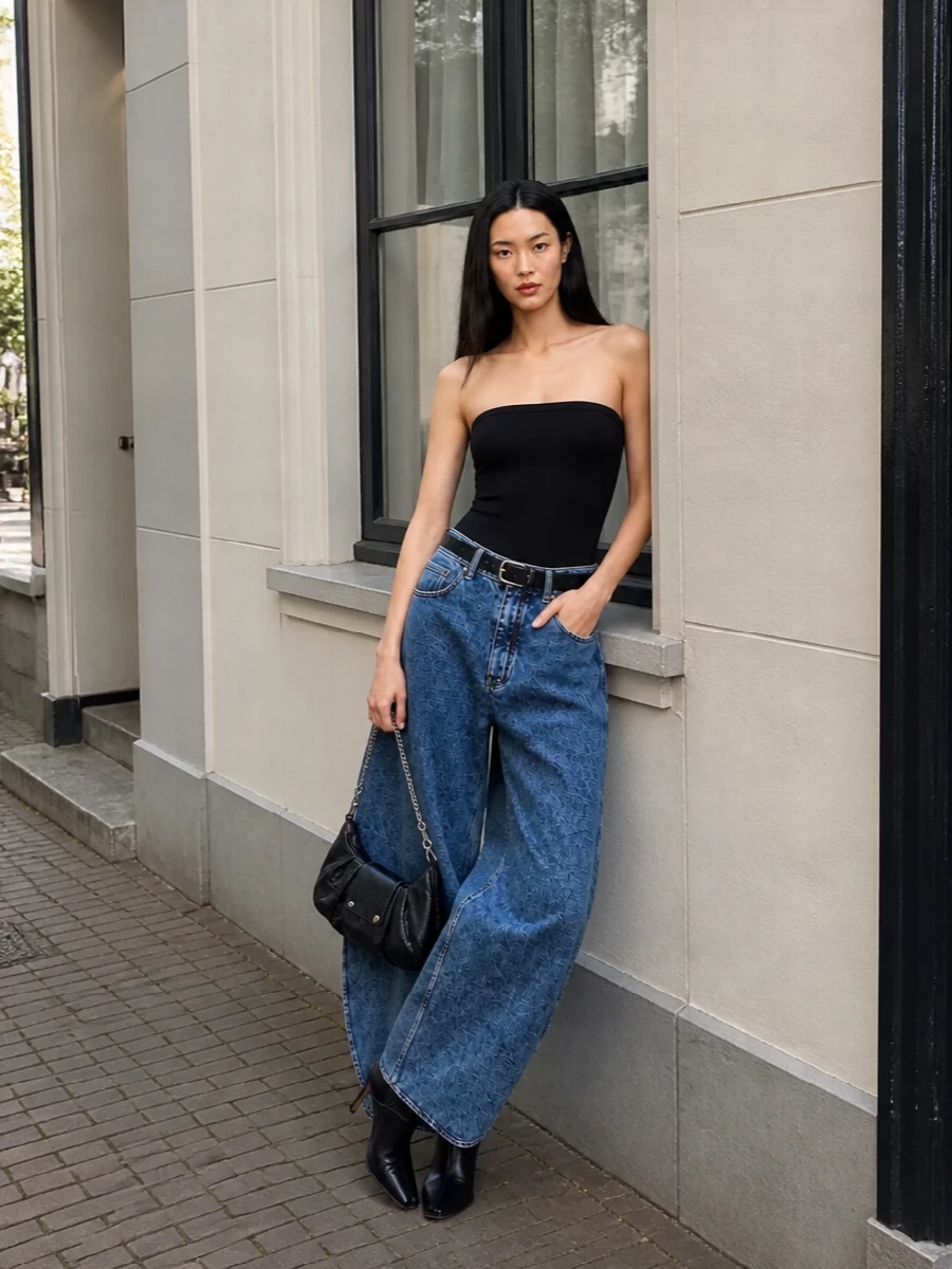 High-rise, ultra-wide Alaïa denim is anchored by a jet-black, strapless bodysuit, creating a sharp hourglass focus above those sculptural legs. Square-toe leather mules and a crinkled black chain baguette push the look firmly into nocturnal, gallery-opening territory, while a slim black belt crisps up the waist so the whole ensemble reads deliberate, architectural and unmistakably luxe.