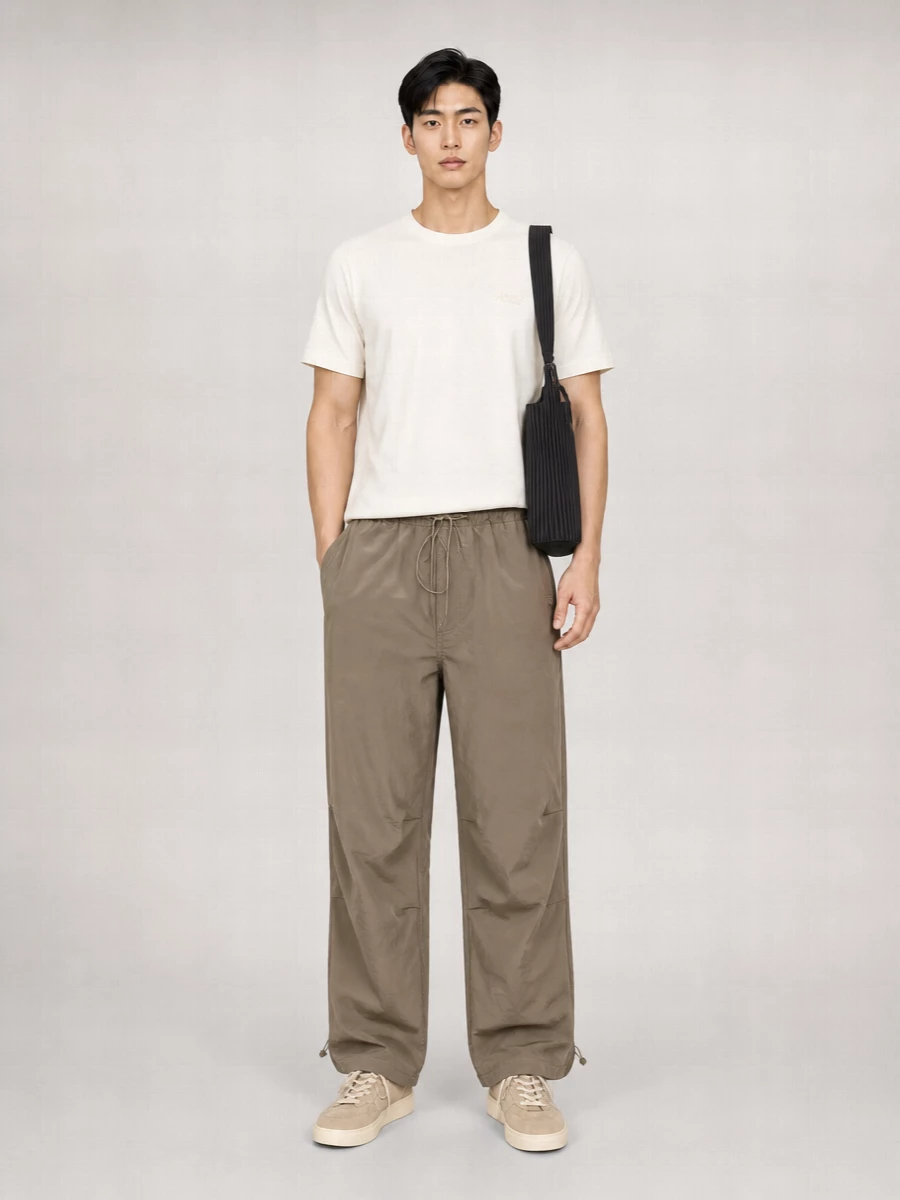 Taupe technical trousers set a relaxed, wide-leg base that feels built for movement, grounded by sand suede sneakers in the same warm spectrum. An off-white tee keeps the upper half clean and bright, while a slim grommeted belt and sculptural black tote introduce subtle structure and texture. The result is a quietly sporty city uniform that feels easy for travel, coffee runs or all-day wear without ever reading sloppy.