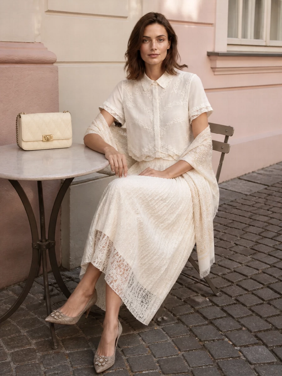 An all-ivory play on texture, this pairing layers sheer lace on lace for a soft, romantic column that still reads polished. The pleated midi balances the cropped shirt, while lace-detailed pumps and a quilted shoulder bag keep the mood quietly luxurious for summer dinners or garden parties. A tonal stole brings just enough extra softness without disturbing the restraint of the palette.