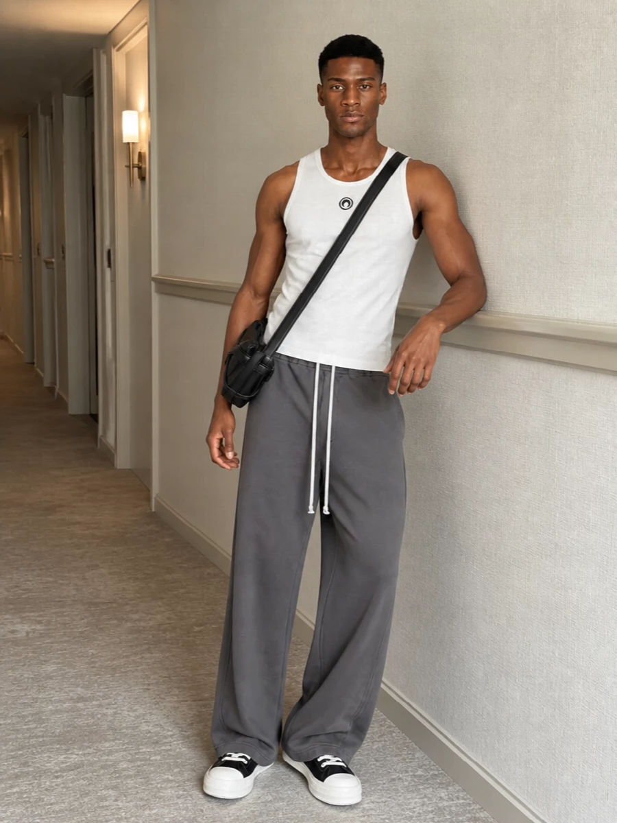 A slim, logo-hit white tank sits against charcoal Rick Owens volume for a clean, street-driven silhouette. DRKSHDW canvas sneakers and a structured McQueen crossbody keep the palette monochrome and the energy technical but relaxed. It feels like modern city sport, more gallery run than gym session.