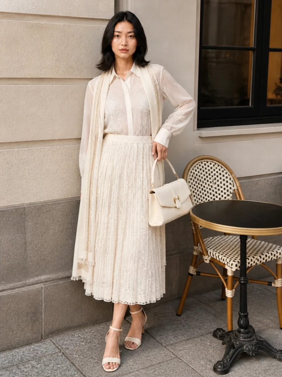 Head‑to‑toe ivory and buttercreams create a soft column of light, with sheer embroidery and pleated lace catching movement rather than attention. The proportions stay long and fluid on top of the high waist, so the midi skirt feels elongating rather than heavy. Barely‑there cream sandals and a structured ivory bag keep the finish polished for summer soirées and candlelit dinners.