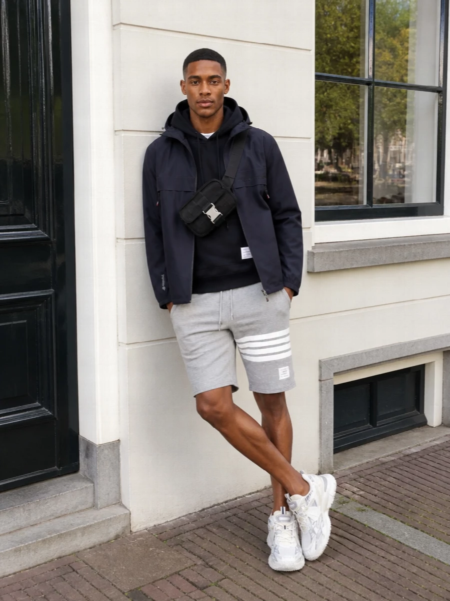 Moncler’s navy shell sits over a deep Thom Browne hoodie and heather-grey jersey shorts for a technical city uniform that still feels considered. Tonal Off-White runners and a compact Gucci belt bag keep the lines clean, the hands free, and the palette quietly graphic from commute to weekend escape.