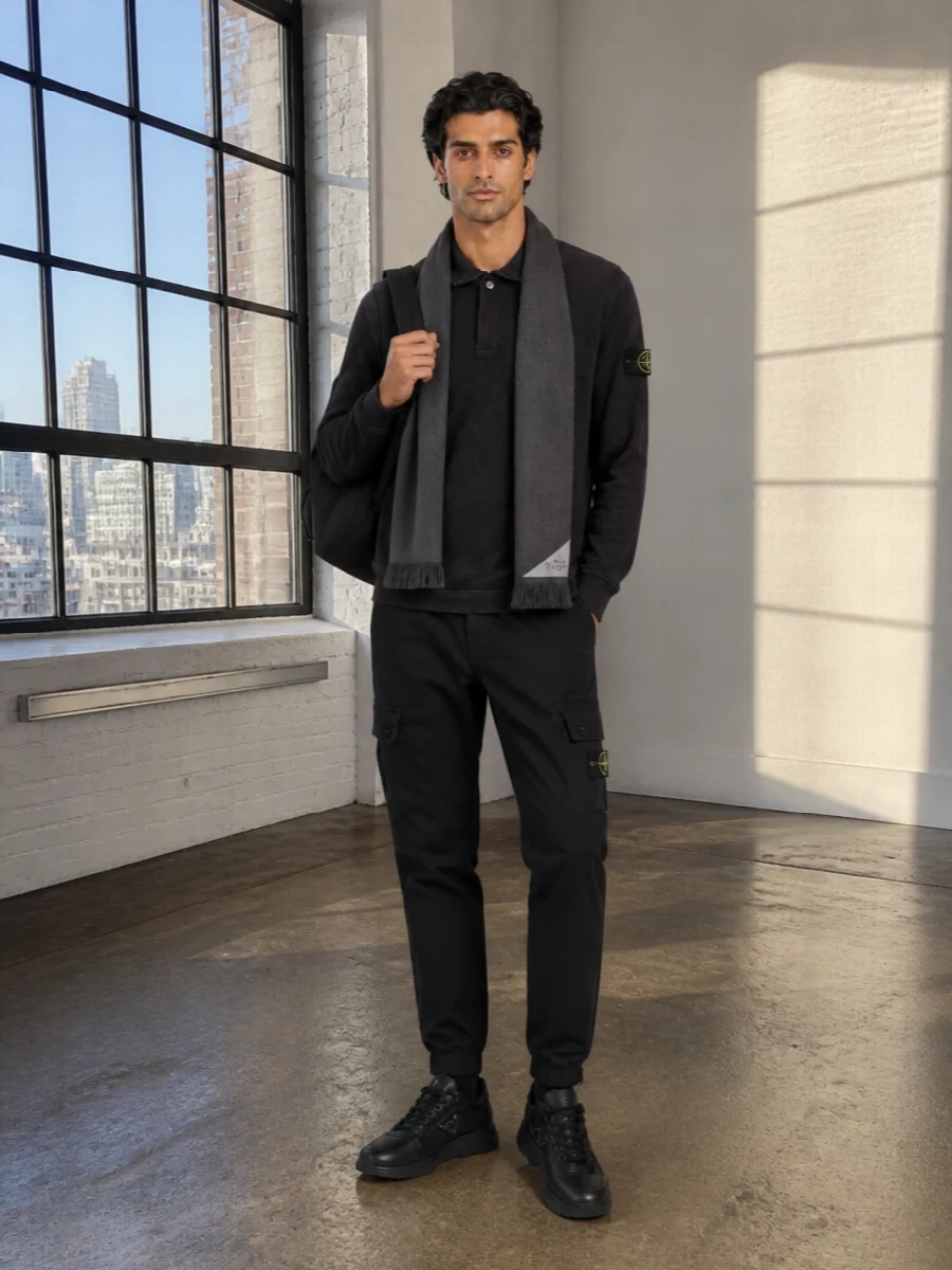 A charcoal Stone Island polo sits over black technical cargo joggers for a clean, street-ready base in a tight monochrome palette. All-black Prada sneakers and a matching nylon backpack sharpen the mood, keeping everything low-key but unmistakably premium. A charcoal scarf finishes the winter story with texture and depth while staying firmly in the minimalist lane.