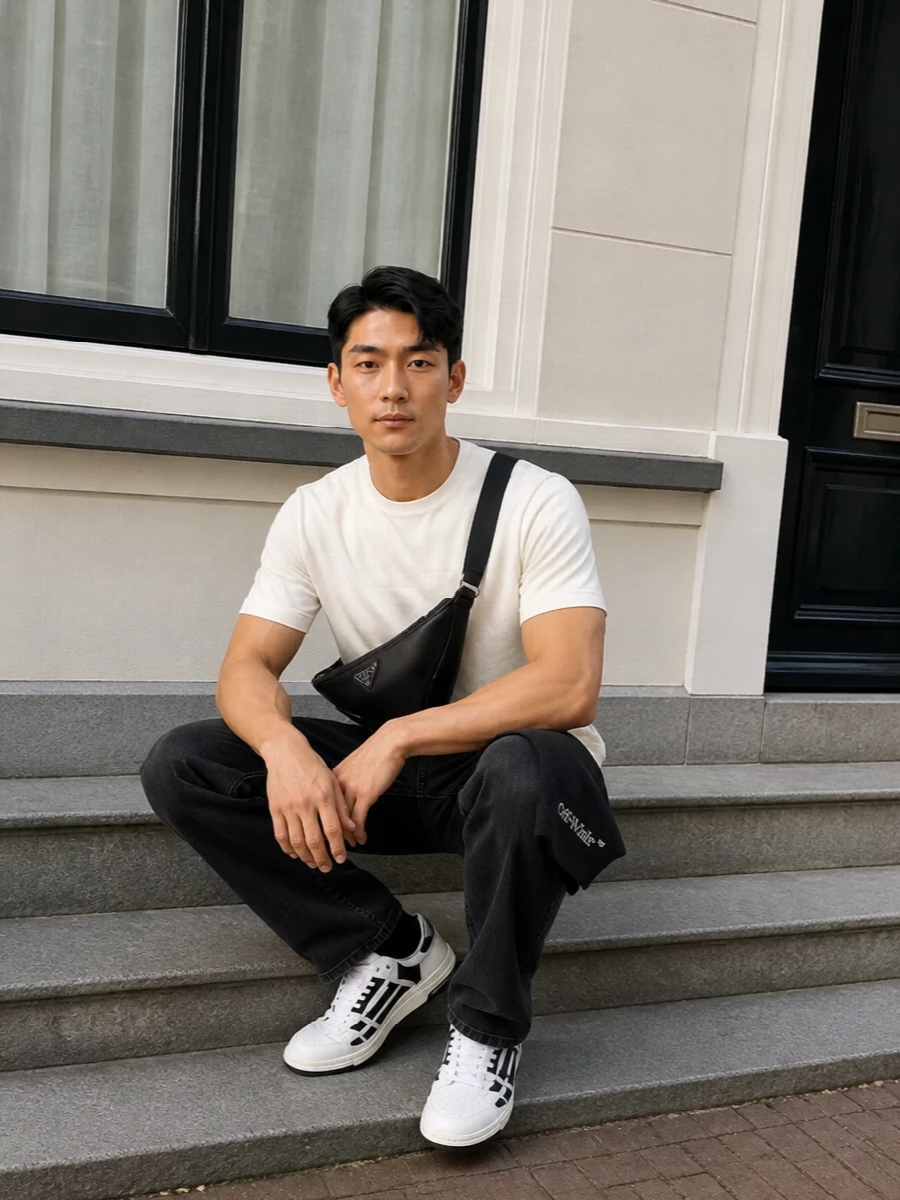 An off-white Amiri tee sits against washed-black straight denim for a clean, graphic monochrome base. Black and white Amiri sneakers echo the contrast, keeping the line relaxed but intentional. A sharp Prada crossbody and discreet logo scarf finish it with city-ready polish that still reads pure streetwear.