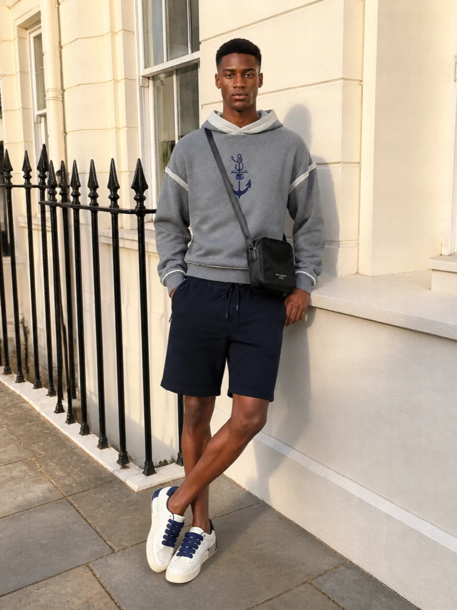Slate grey, navy and off-white stay tight and graphic, so the anchor hoodie feels intentional rather than casual. Relaxed navy shorts and padded sneakers keep the proportions easy, while the compact crossbody adds just enough urban edge. It is a clean, logo-driven streetwear outfit built for warm off-duty days and low-key travel.