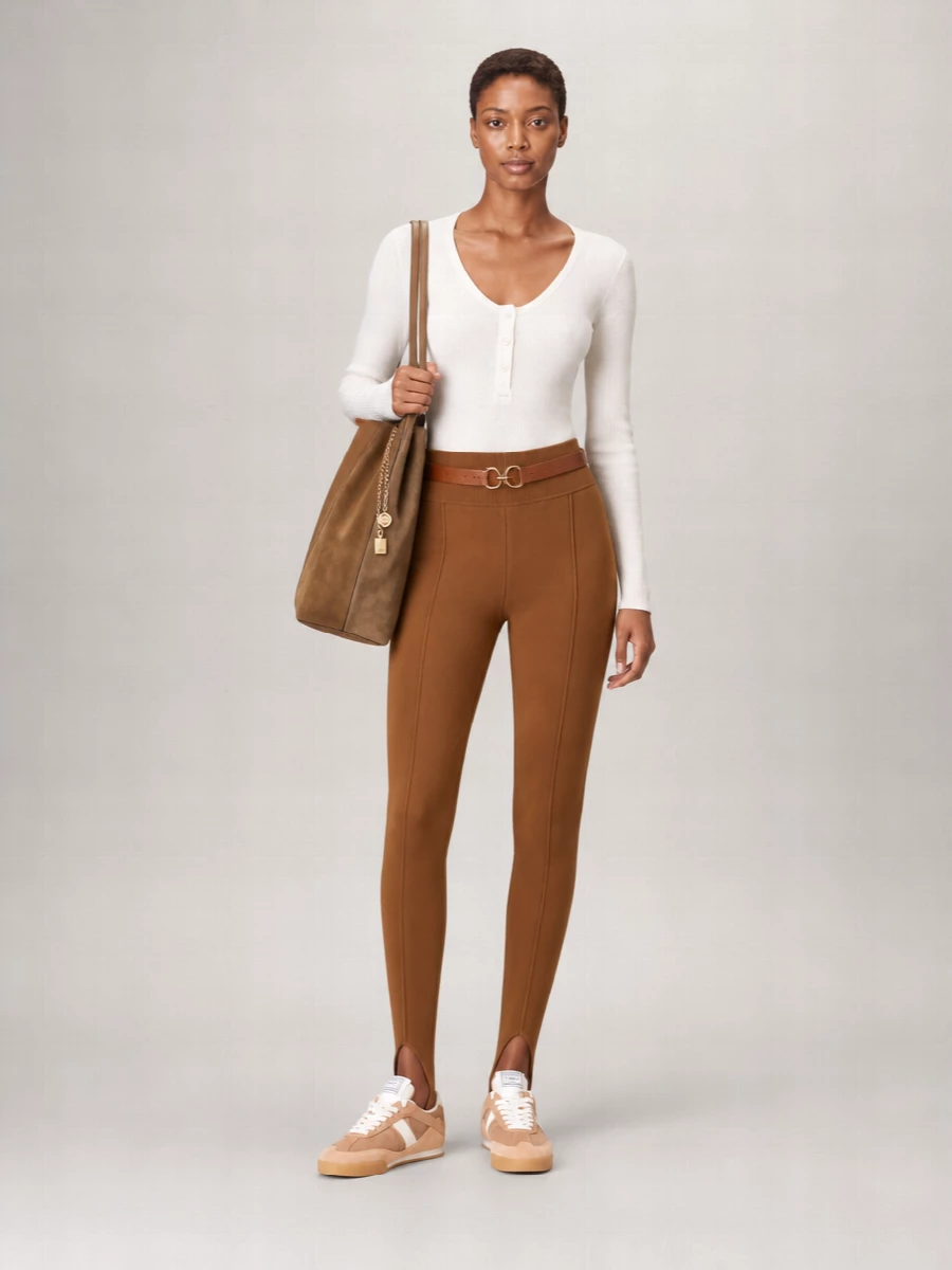 Caramel stirrup leggings meet a slim ivory knit for a clean, body-tracing base that feels relaxed yet intentional. Camel-toned sneakers and a spacious tan tote keep everything tonal and practical, while a cognac belt quietly sharpens the waistline. The result is a softly tailored, neutral uniform that works for travel, off-duty meetings, or polished weekends in the city.