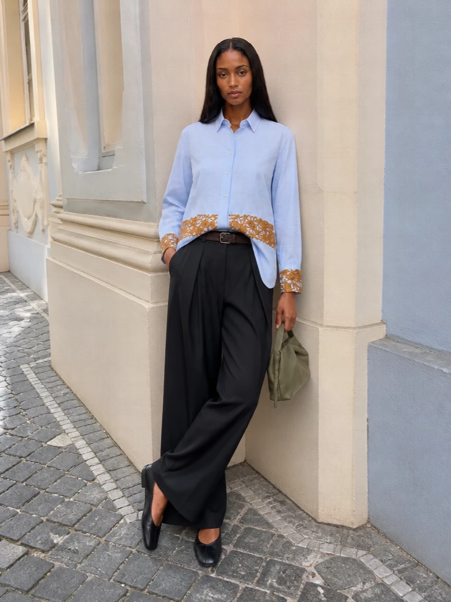 A pale blue embroidered shirt meets fluid charcoal tailoring for a long, calm line that feels precise without stiffness. The square toe flat and slim belt keep the volume grounded and intentional, while the sage clutch adds a muted, intelligent note of color. It reads like modern day dressing for someone who prefers nuance over noise.