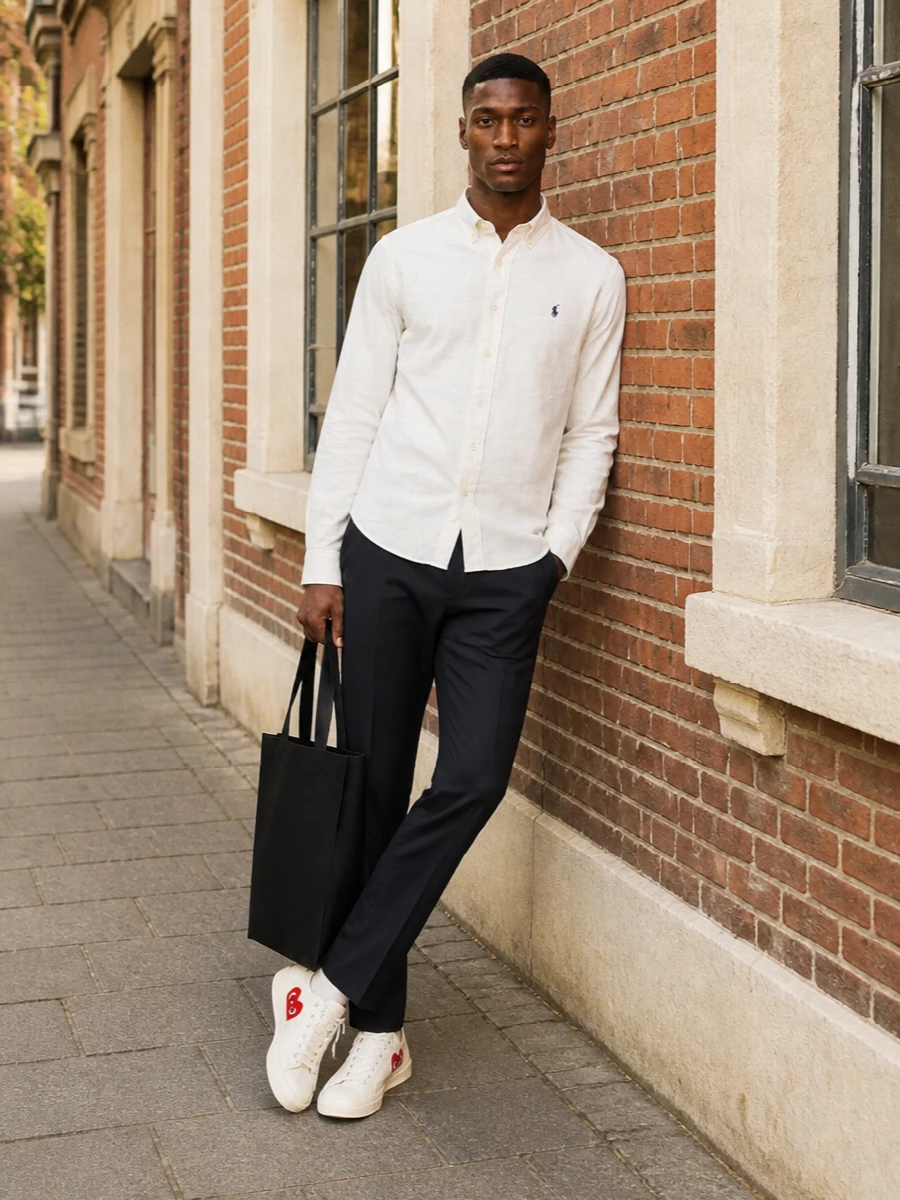 A crisp ivory Oxford shirt is paired with deep charcoal tailored trousers for a sharp, all-season smart-casual base. Off-white canvas sneakers relax the mood without losing polish, while a structured black tote keeps the whole mix clean, functional and city ready.