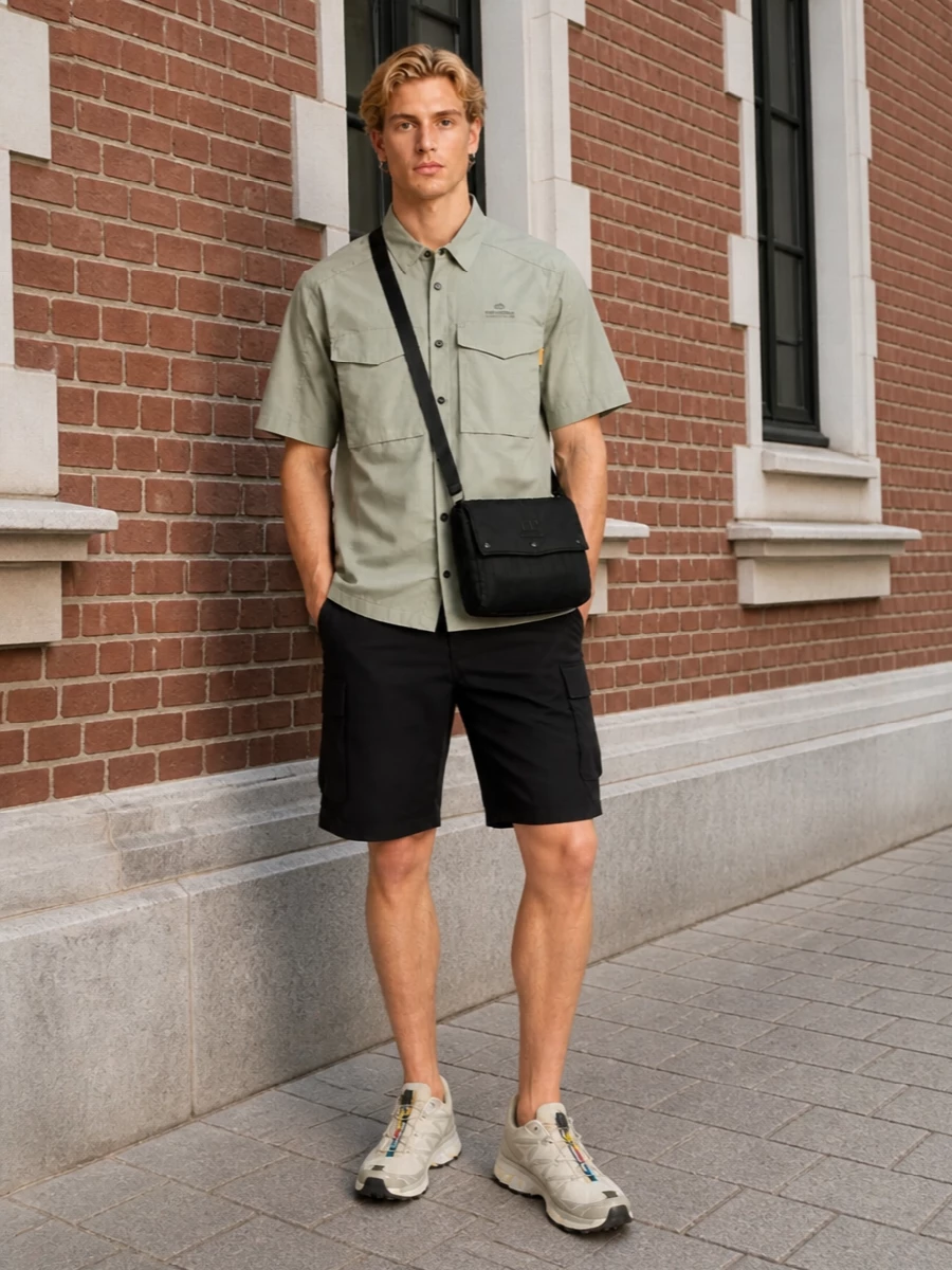 Sage technical shirting sits over matte black cargo shorts for a clean, urban utility base. Neutral trail runners in soft beige keep the palette light while still performance-focused. A compact black crossbody finishes it with practical structure and low-key branding for city days and travel.