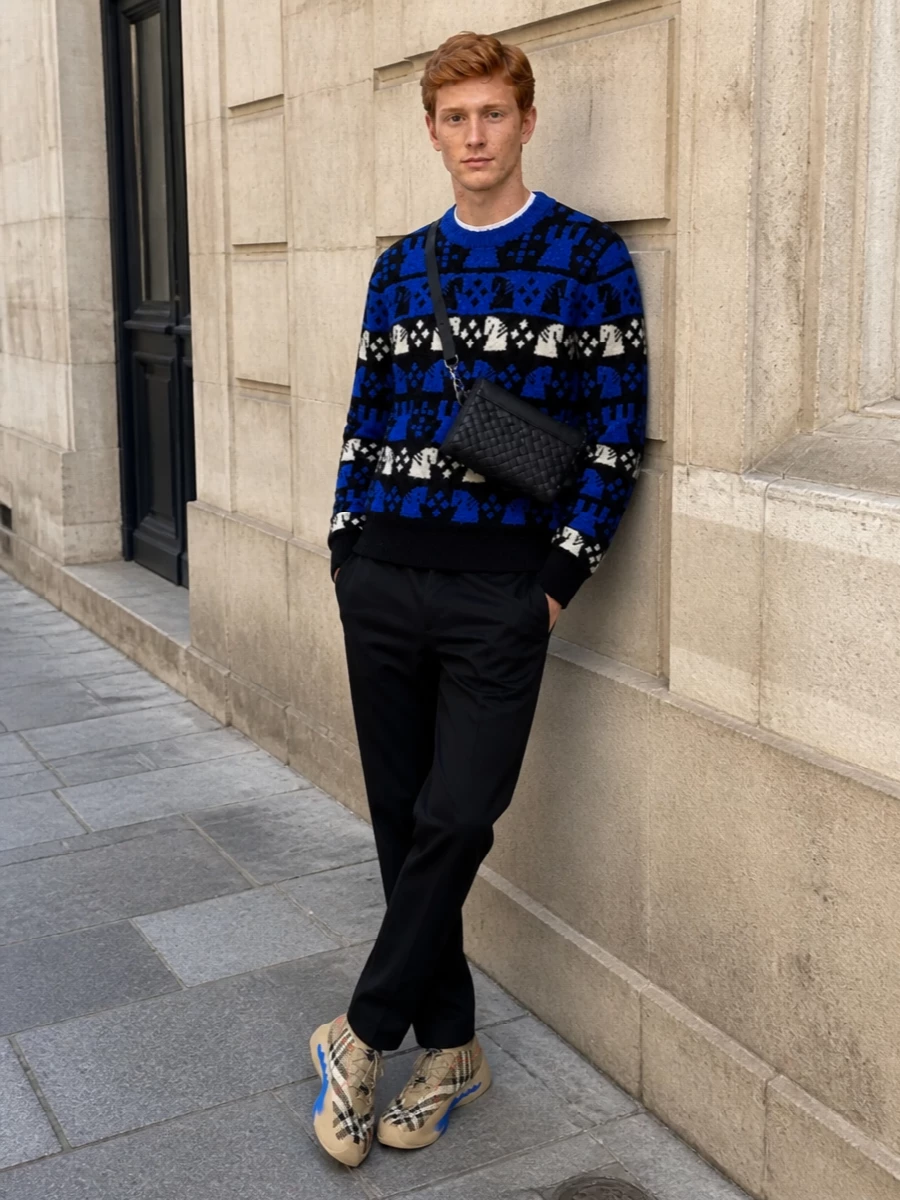 This look lets the sneakers and knit do the talking: the cobalt-and-ivory chess-motif sweater picks up the shoe’s blue sole and graphic patterning, while the black chinos keep the line clean and streamlined. A sleek black Bottega crossbody adds discreet luxury and practicality, polishing off a modern FW city uniform that feels bold yet controlled.