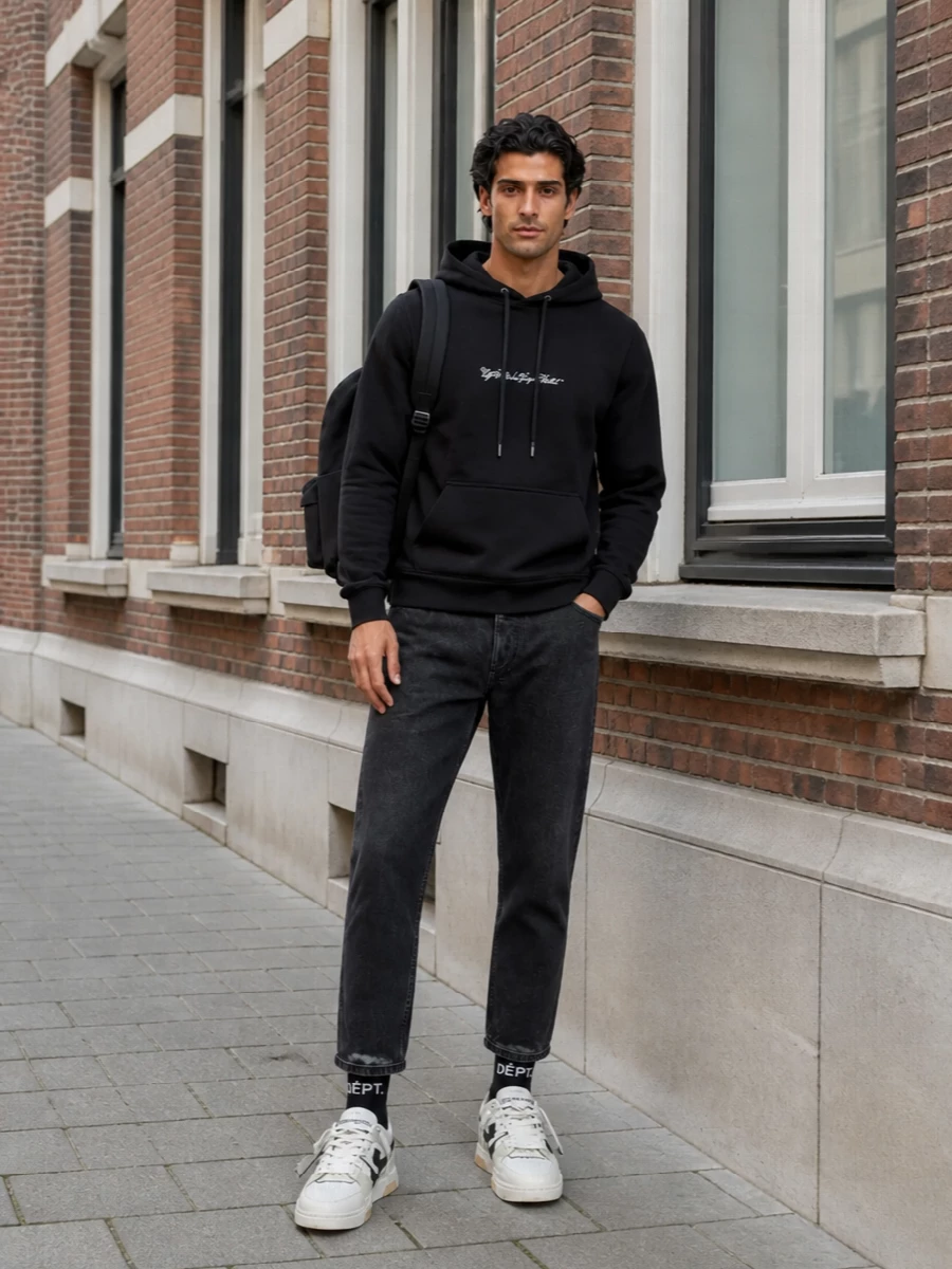 A black Off-White hoodie meets washed black straight-leg denim for a monochrome street take that feels relaxed but intentional. The chunky mesh sneakers lighten the base just enough, while the clean Balenciaga backpack and graphic socks keep the whole outfit reading as modern city uniform rather than gym gear.
