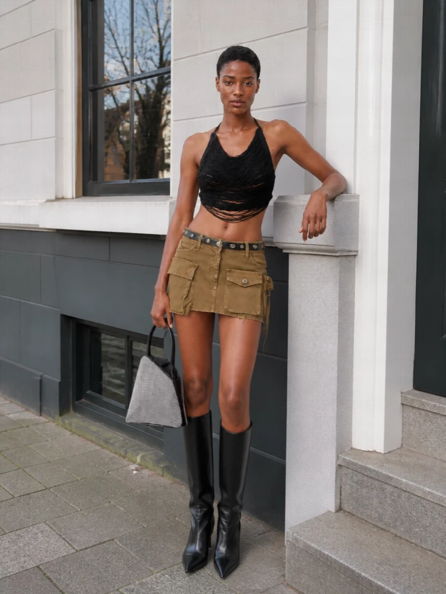 This is a legs-for-days city outfit built around the low-slung khaki cargo mini, sharpened with a black fringe halterneck that keeps everything firmly in after-dark territory. Glossy pointed knee boots extend the line of the leg while the studded belt and crystal bucket bag echo each other in hardware and shine. The result feels like Attico nightlife uniform: utility, body-conscious and unapologetically bold in a grounded khaki and black palette.