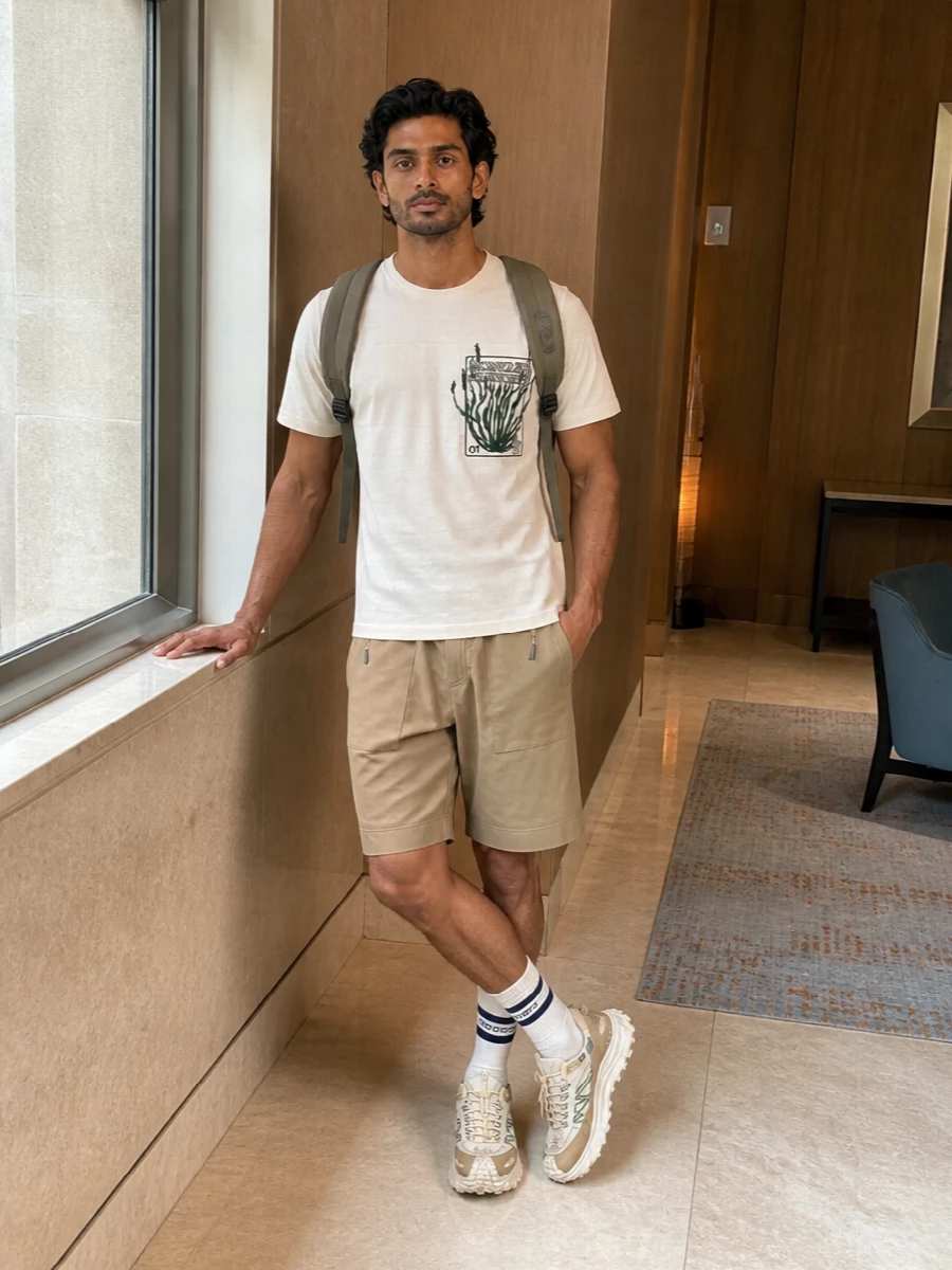 A cream graphic tee meets sand technical cargos for a light, trail-ready city uniform built on clean lines and easy movement. Cream and olive-accented trail sneakers echo the tee’s print, while a taupe technical backpack keeps the palette grounded in earthy neutrals. Off-white striped socks add a quiet sport detail without distracting from the core monochrome and green story.