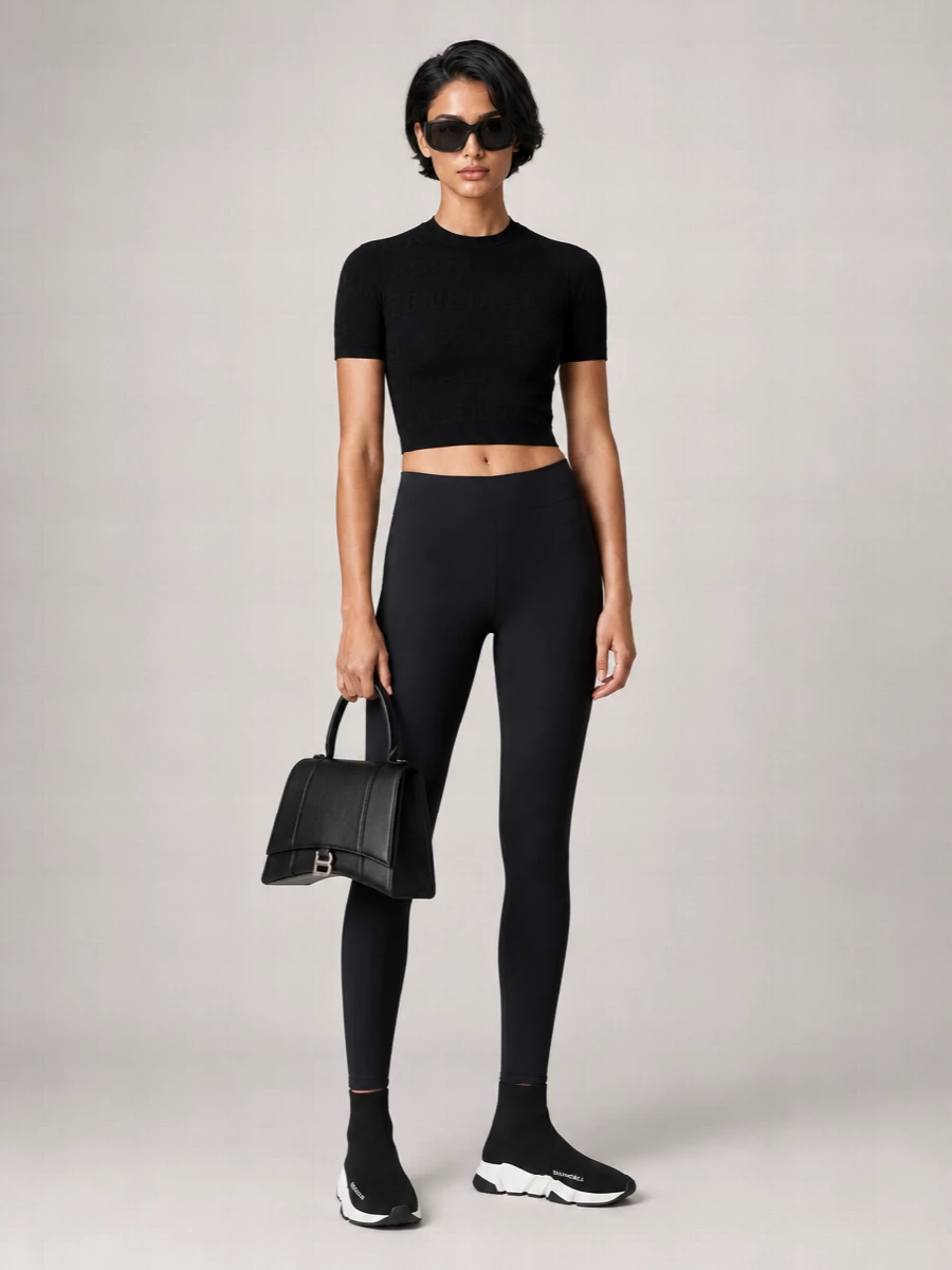 A deep-black Balenciaga column on top and bottom keeps the line clean and fast, with the crop of the tee flashing just enough skin to break up the silhouette. Sock-like sneakers extend that second-skin idea into the footwear, while a structured trapeze bag and sculptural sunglasses sharpen the mood from gym-adjacent to city-ready.
